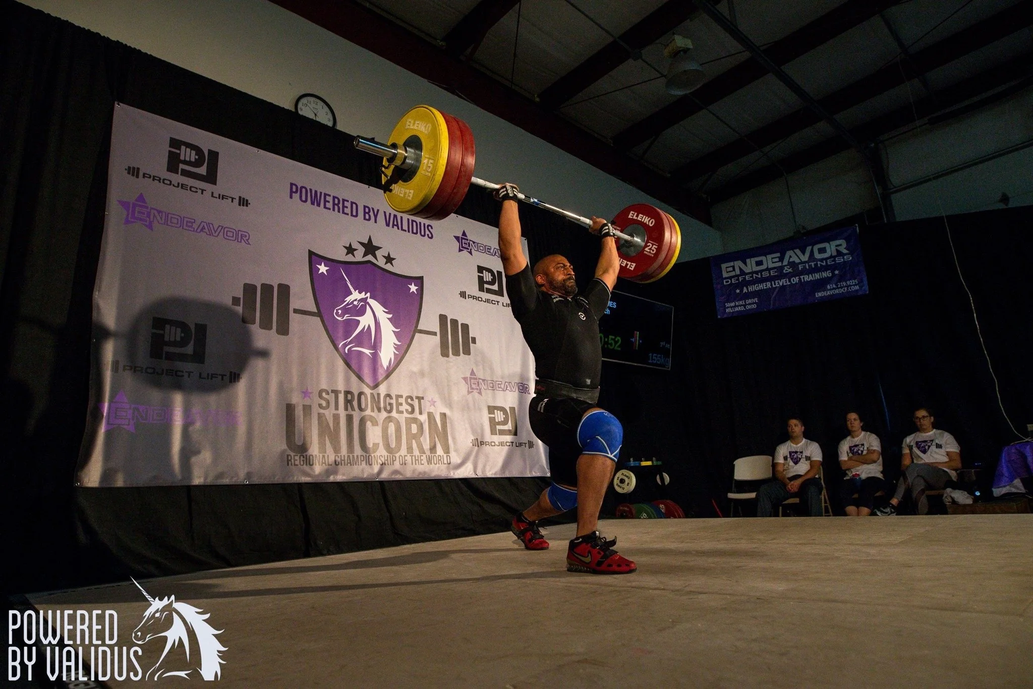 A male powerlifter in black clothing and red shoes lifts a barbell overhead during a competition, with a banner in the background displaying event logos and the phrase 'Strongest Unicorn'.