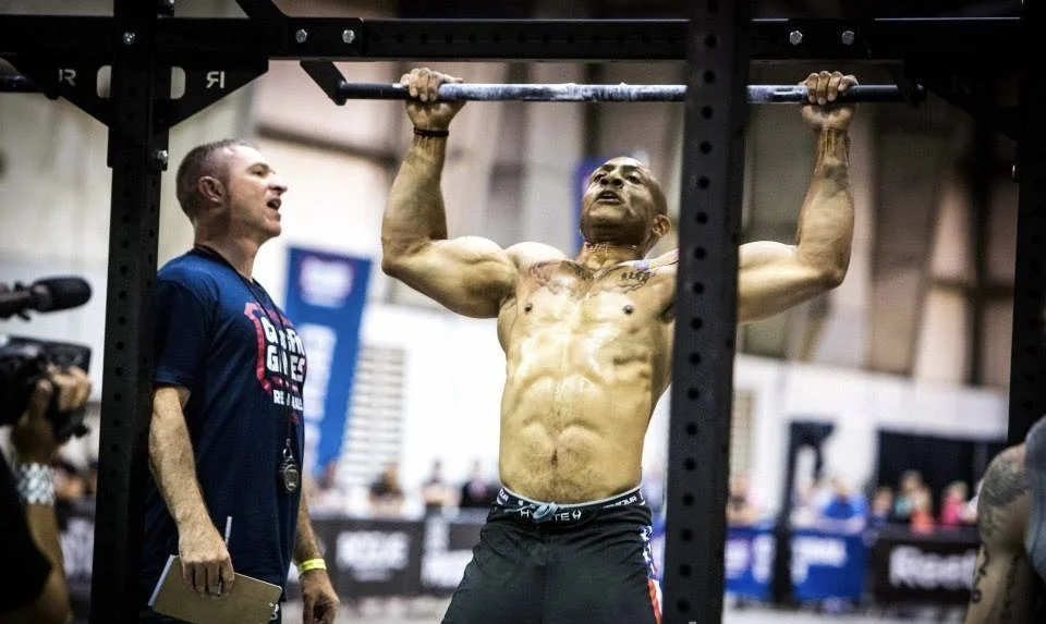A muscular man performing a pull-up on a bar at a fitness competition, with a coach or judge monitoring, other people in the background, and a camera recording.