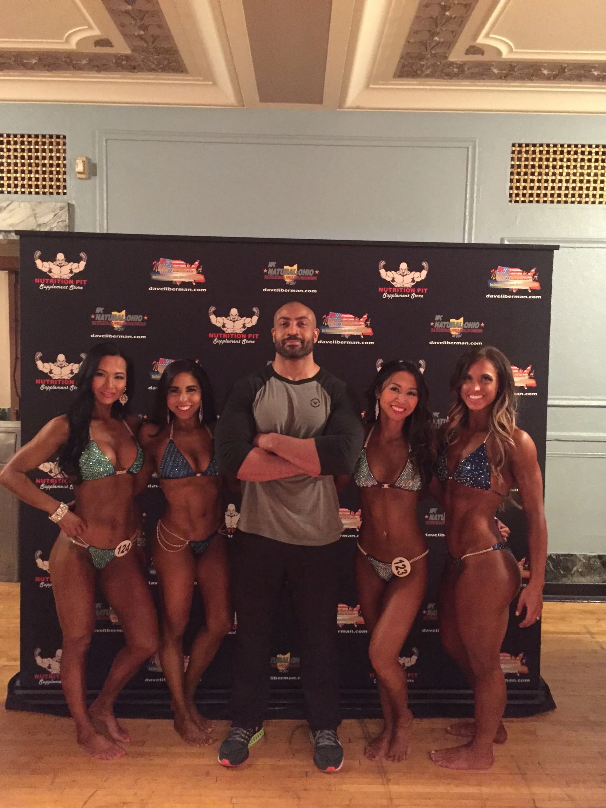 Group of five people, four women in bikinis and one man in casual clothing, standing on a wooden floor in front of a black backdrop with logos for 'Nutrition Pit', 'daveliberma.com', and 'In Natural Ohio'.
