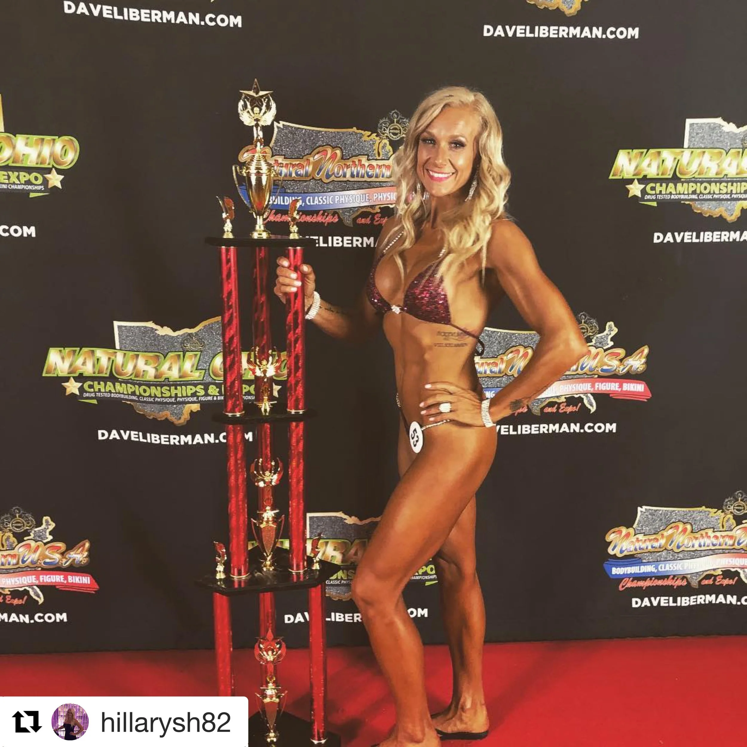 A woman in a bikini standing on a red carpet, holding a tall red and gold trophy, at a bodybuilding competition with a backdrop featuring event logos and website.