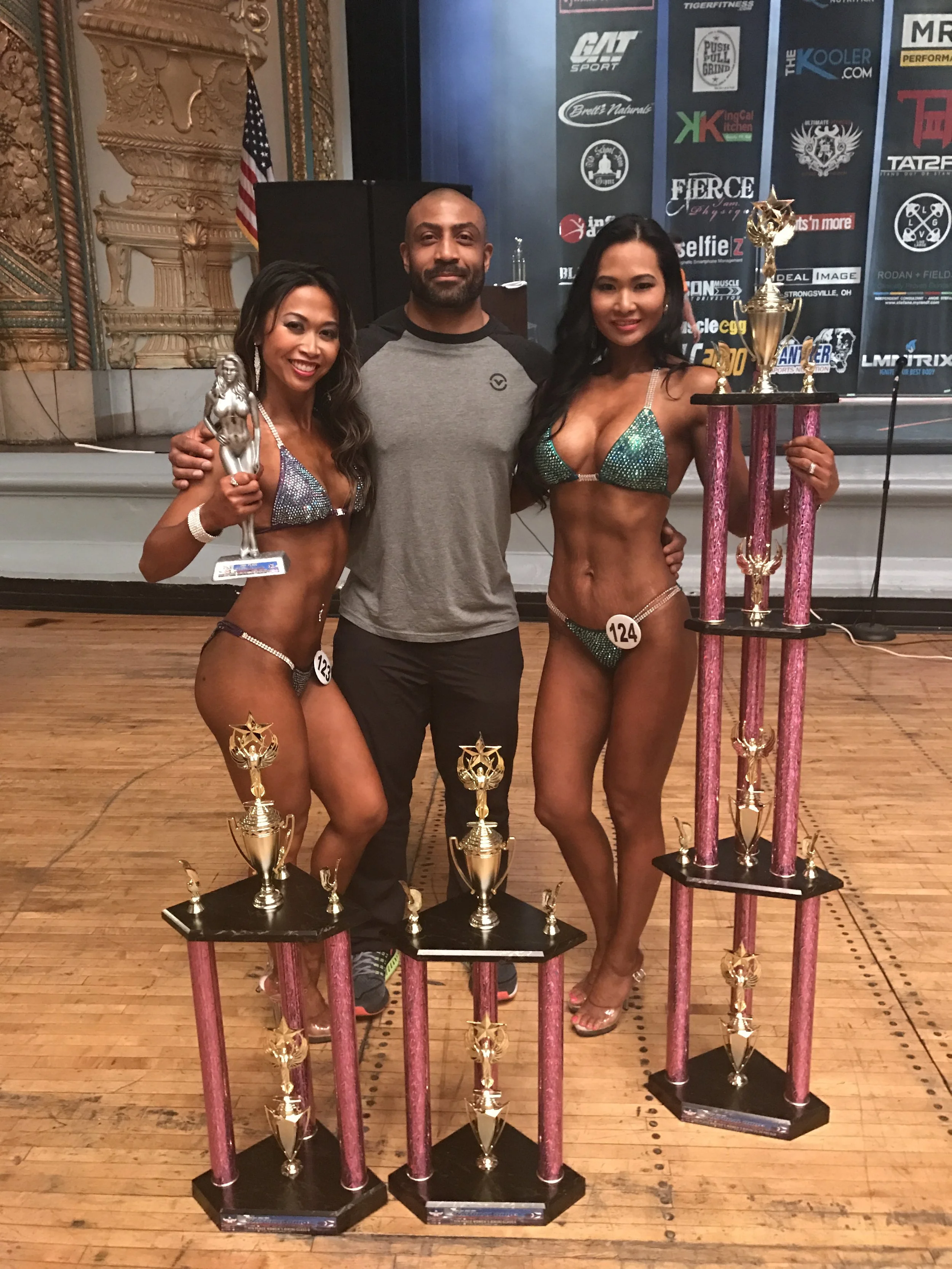 Two female bikini bodybuilders in the bikini competition with trophies, standing with a man in the center. They are on a stage with a backdrop showing numerous sponsor logos. One woman holds a silver trophy, and the other holds a large multi-tiered t