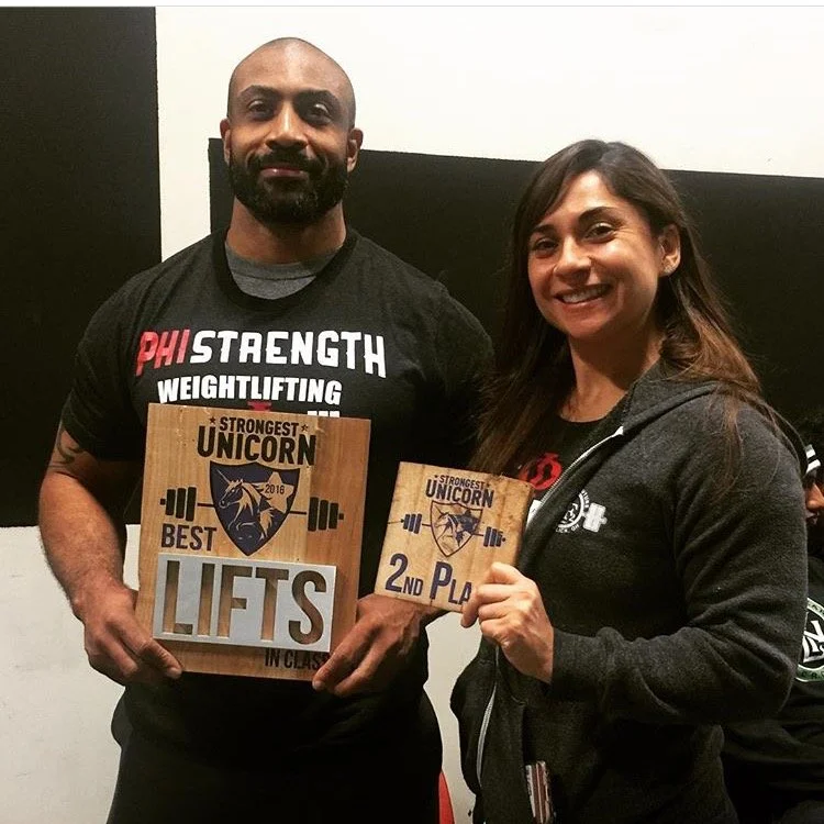 A man and woman posing indoors, holding plaques and medals from a powerlifting competition. The man is wearing a black shirt with sports-related text, and the woman is wearing a black hoodie with a logo, both smiling.