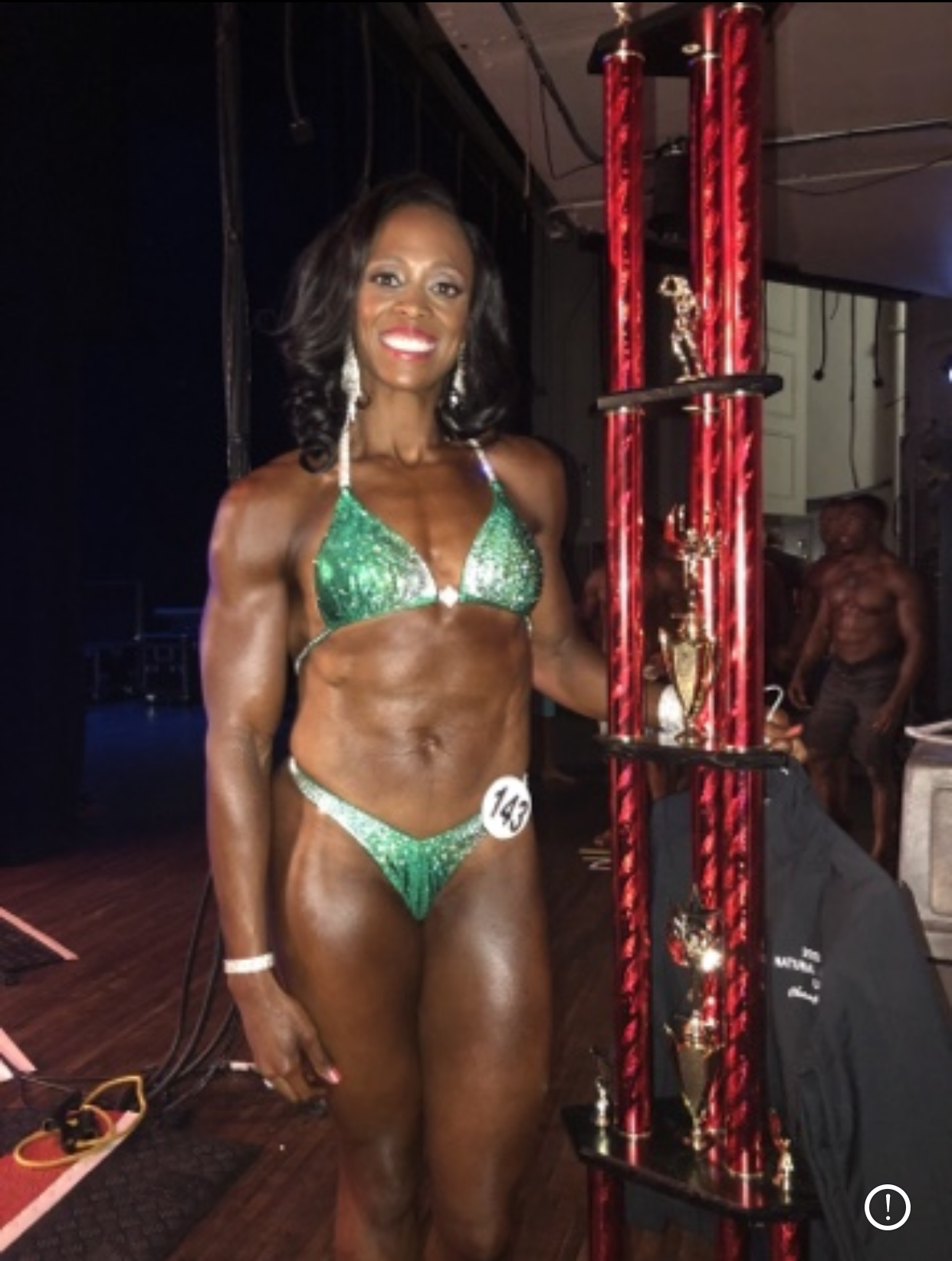 A woman in a green bikini with the number 143 at a bodybuilding competition, standing next to a tall red and gold trophy.