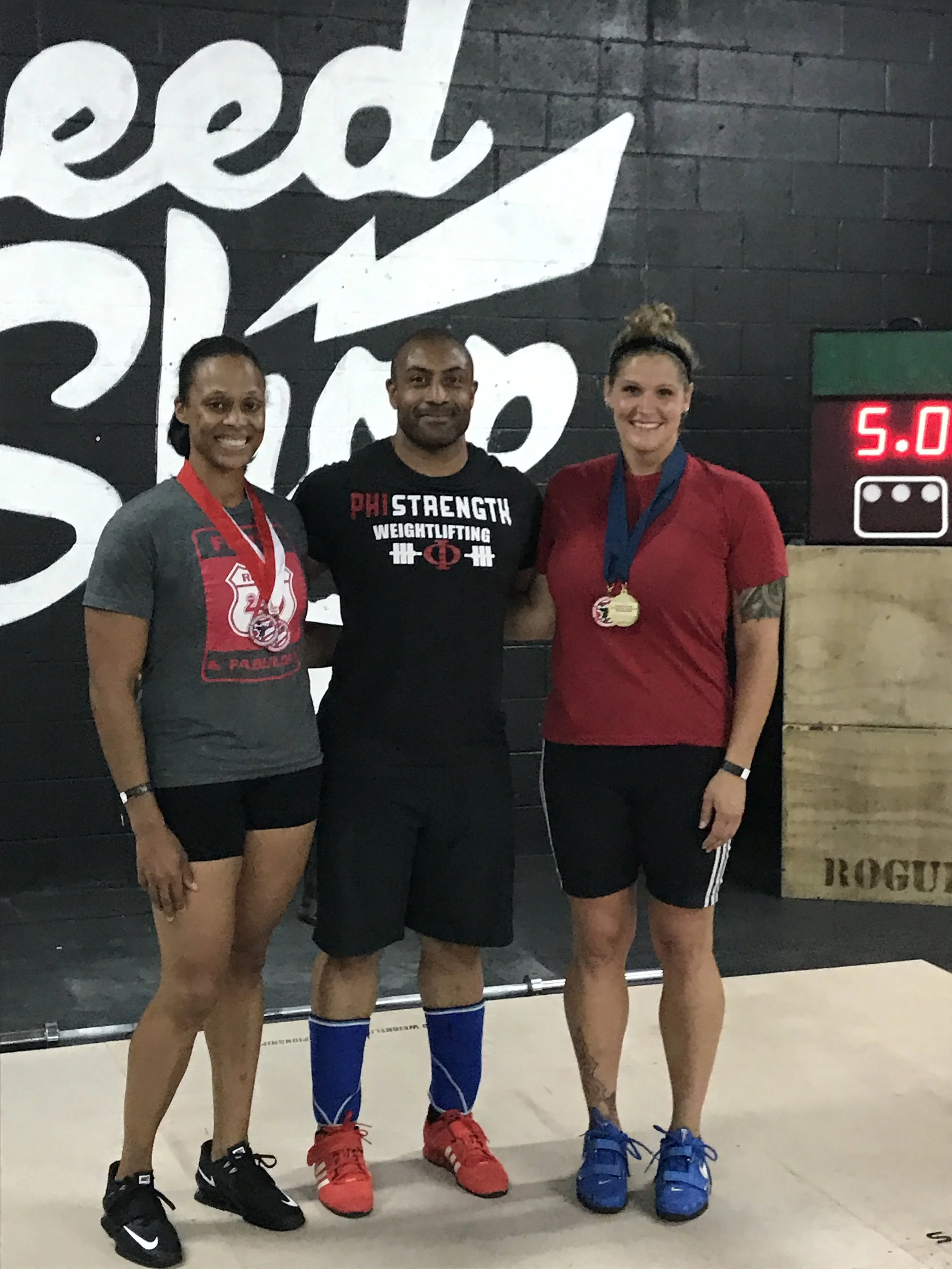 Three women and one man standing together inside a gym, posing for a photo after a weightlifting competition. The women are wearing medals around their necks, and the woman on the right has a tattoo on her arm. The man is wearing a shirt that reads '