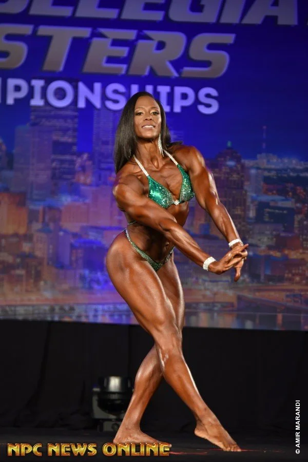 A female bodybuilder on stage in a green bikini with sparkling details, posing to showcase her muscular physique during a bodybuilding competition, with a cityscape backdrop and the words 'NPC Championships' in the background.