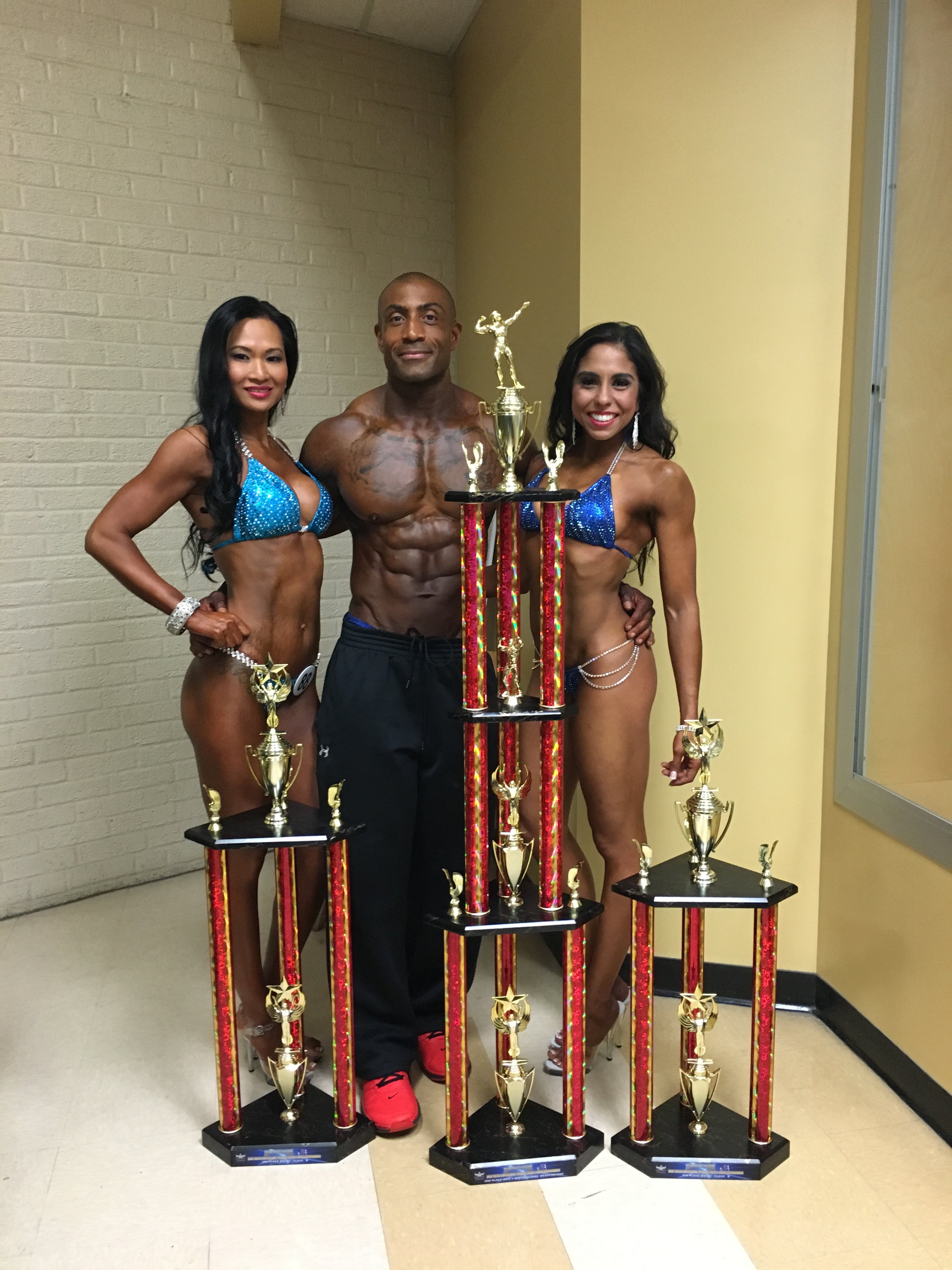 Three muscular individuals in bikinis and athletic shorts standing behind tall trophies, posing in an indoor setting.