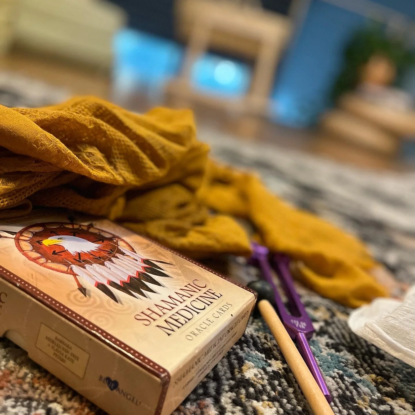 A box of Shamanic Medicine oracle cards, a yellow fabric cloth, a purple crochet hook, a wooden stick, a white tissue, and a black bead are laid out on a patterned carpet with a blurred background of furniture and a potted plant.