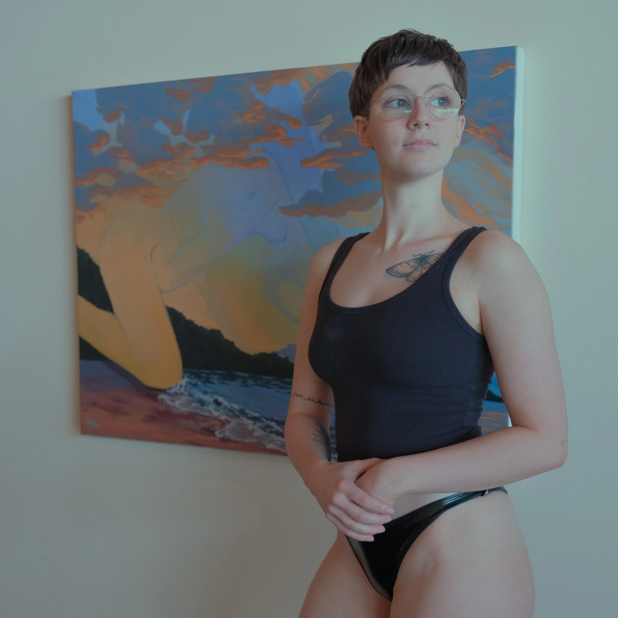 A woman with short dark hair, wearing glasses, a black tank top, and black underwear, standing in front of a colorful landscape painting of a sunset over water and mountains.