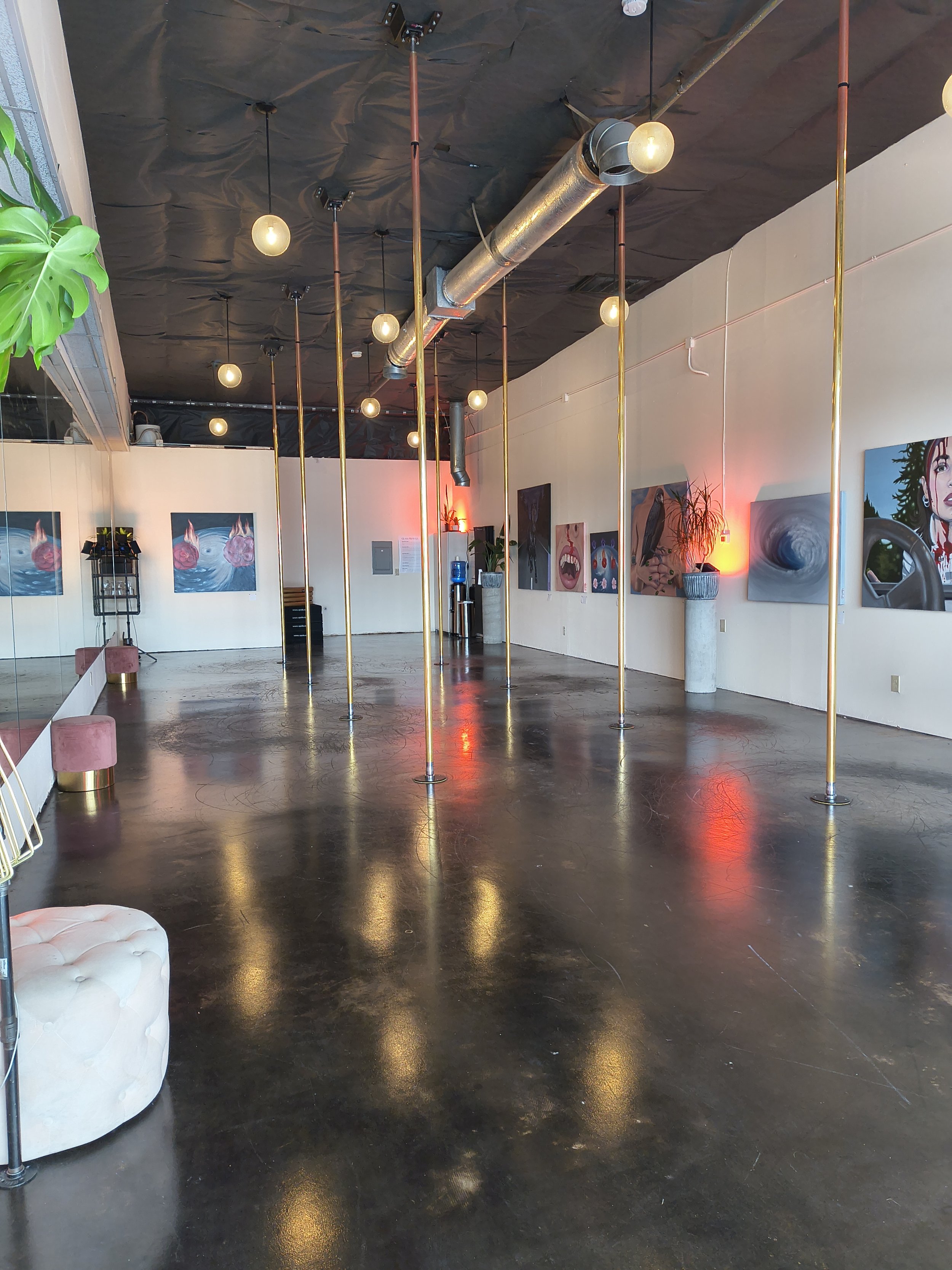 An empty dance studio or event space with black polished concrete floors, white walls decorated with colorful artwork, a ceiling with black insulation, and decorative globe lights hanging from the ceiling. There are also several gold-colored poles arranged in a circle in the center of the room.