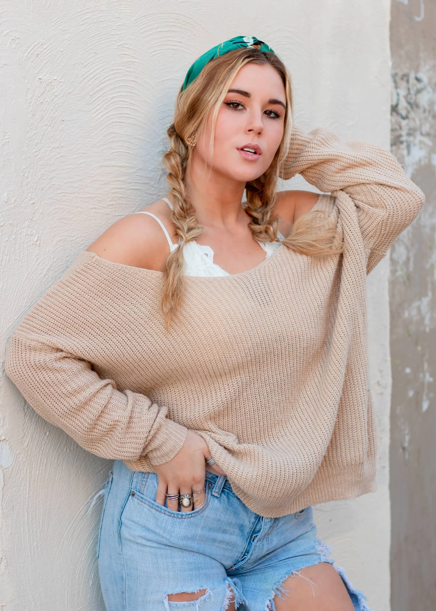A young woman with blonde hair in braids wearing a teal headband, off-shoulder beige sweater, white lace top, and ripped denim shorts, posing against a textured wall.