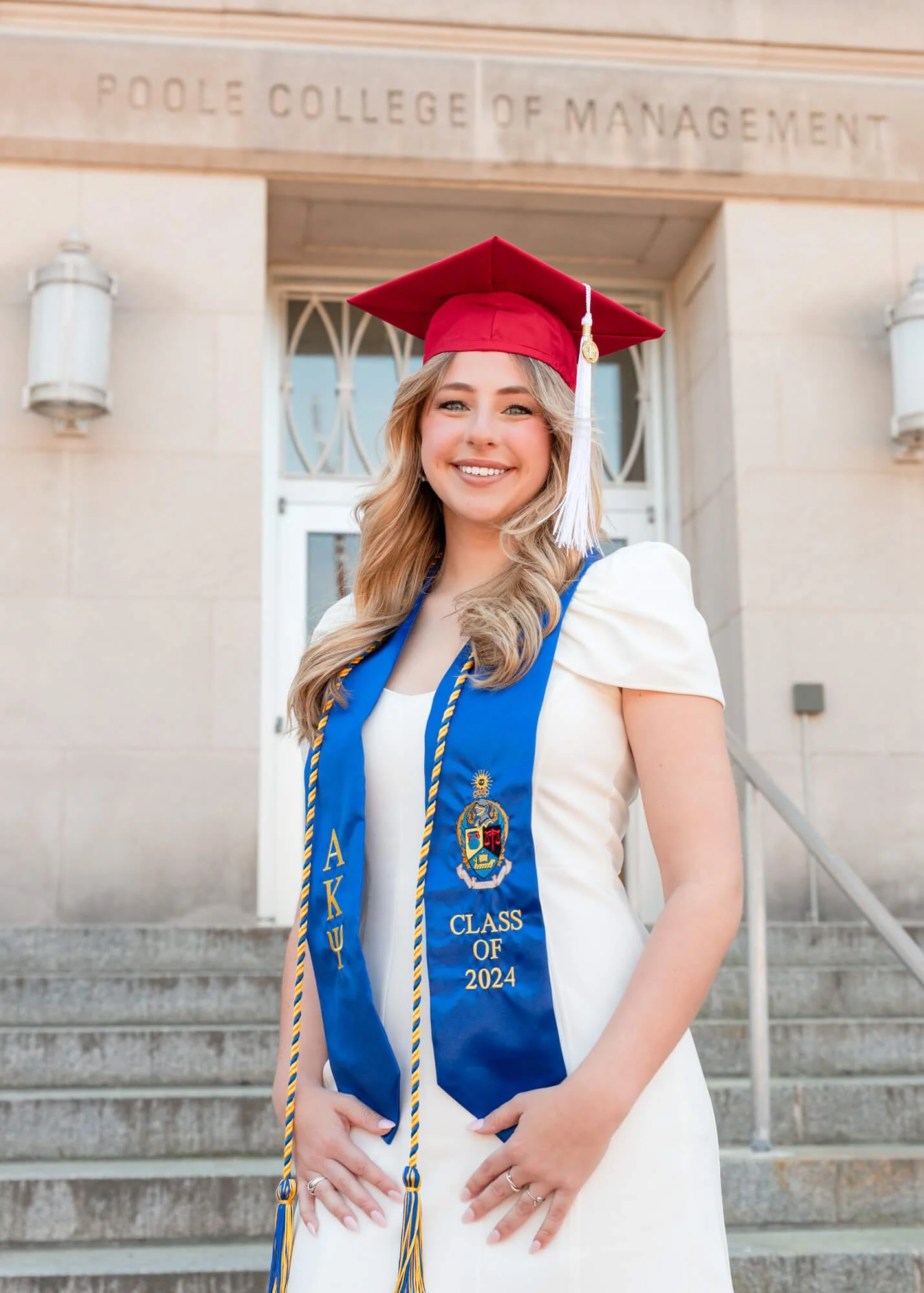 A young woman in a white dress and graduation cap standing on steps in front of a building with the sign 'Poole College of Management'. She is smiling and wearing a blue graduation stole with a crest, gold embroidery, and the text 'AKΨ Class of 2024'