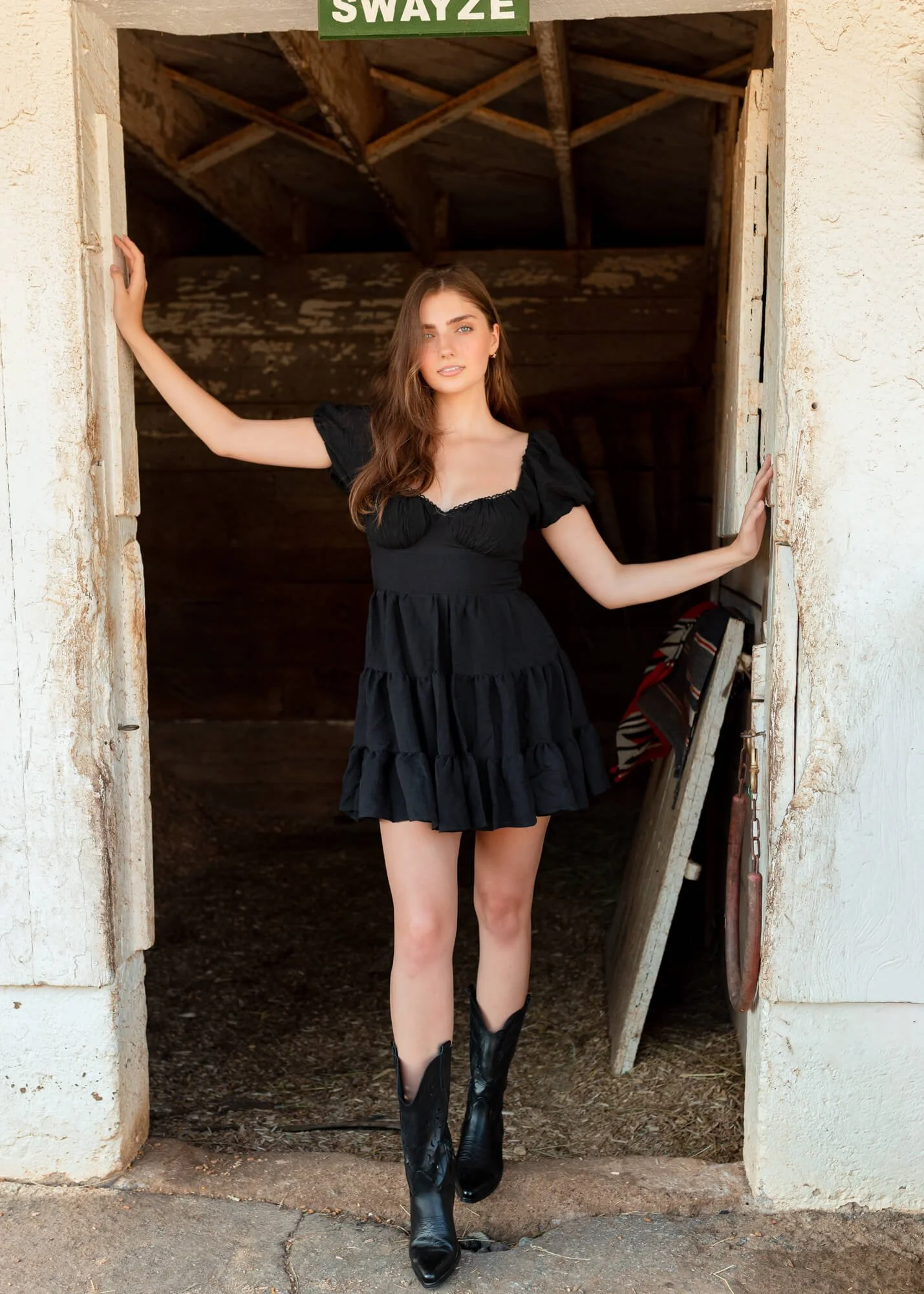 A young woman in a black dress and cowboy boots standing in a barn doorway, with her arms resting on the doorframe.
