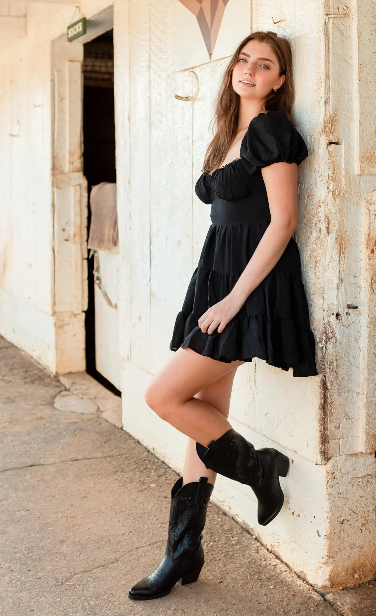 A young woman with long brown hair wearing a black dress with puffed sleeves and cowboy boots, leaning against a rustic beige wall outside an old building.