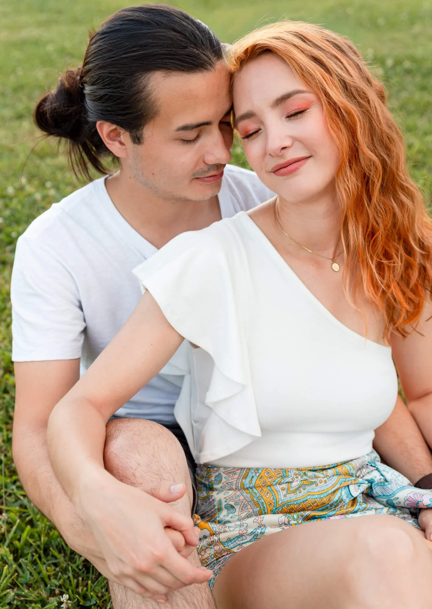 A man and woman sitting on grass with eyes closed, embracing each other, with the woman wearing patterned shorts and a white top.
