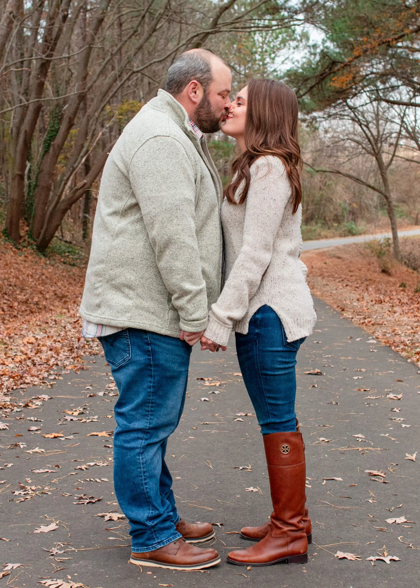 A couple kissing and holding hands outdoors on a fall day, with trees and fallen leaves around them.