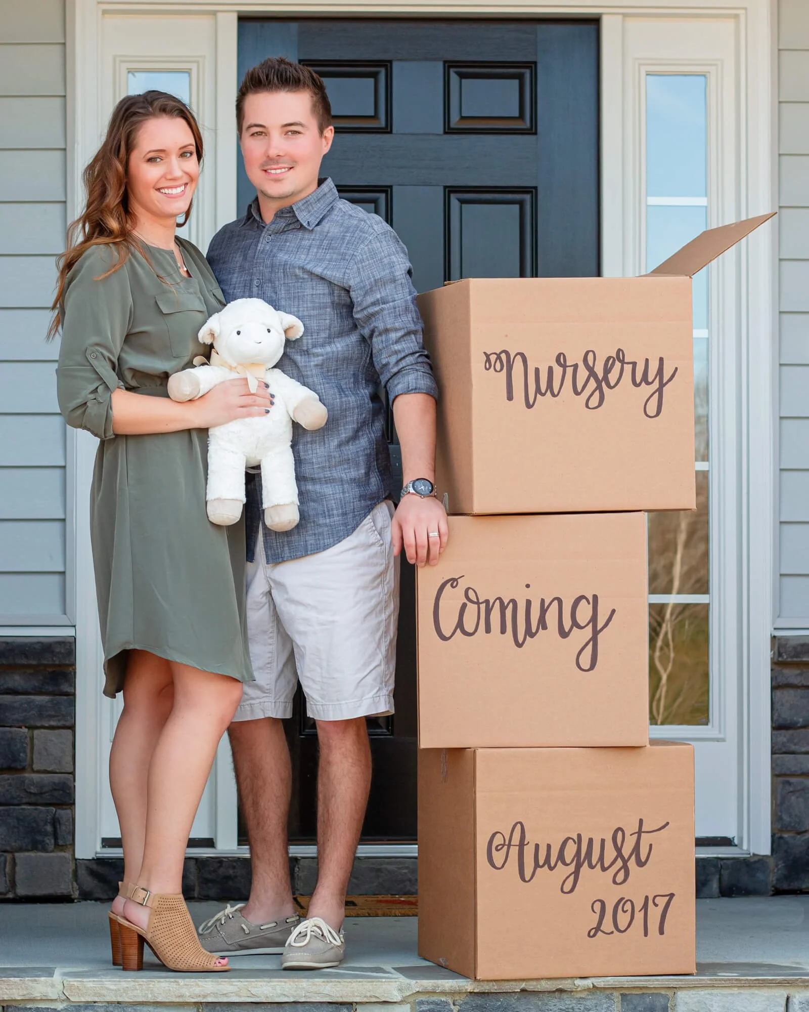 A young couple standing in front of a house with cardboard boxes labeled "Nursery" and "Coming August 2017", holding a stuffed sheep toy, smiling, as they prepare for a new baby.