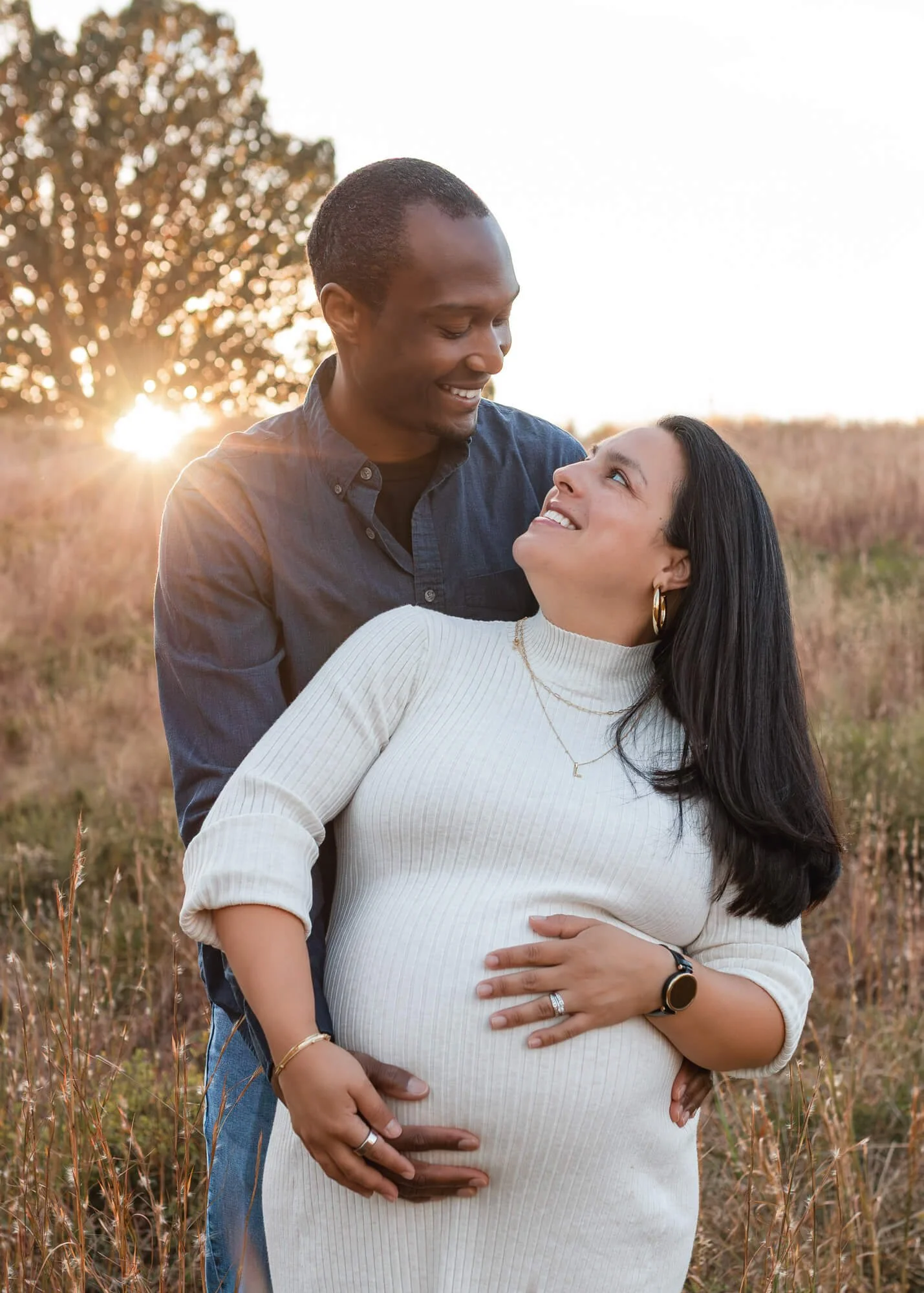 A couple standing in a field at sunset, with the man smiling down at the pregnant woman, who is looking up at him.