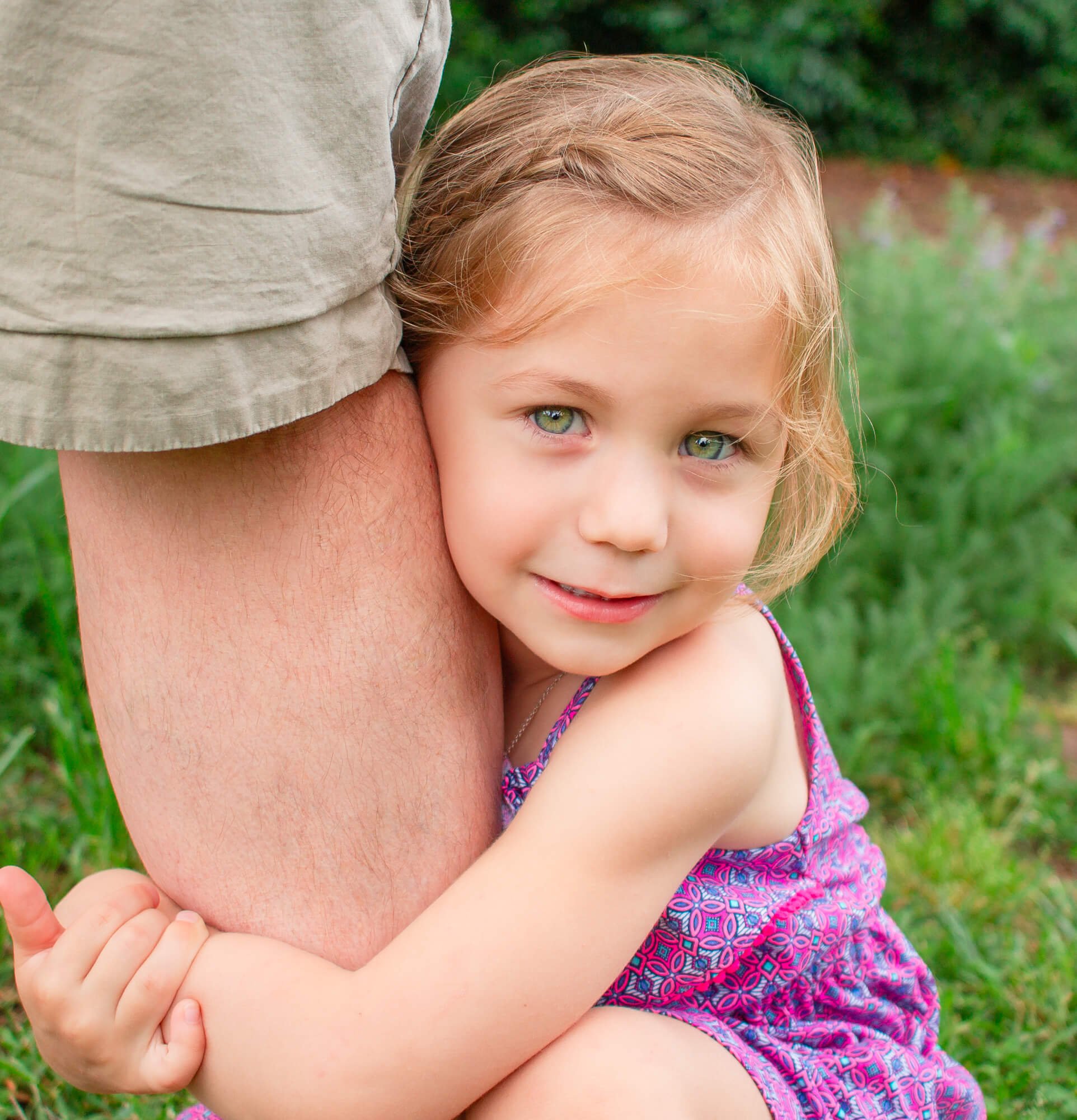 A young girl with blonde hair and green eyes hugging an adult's arm outdoors on green grass, wearing a colorful dress with pink and purple patterns.