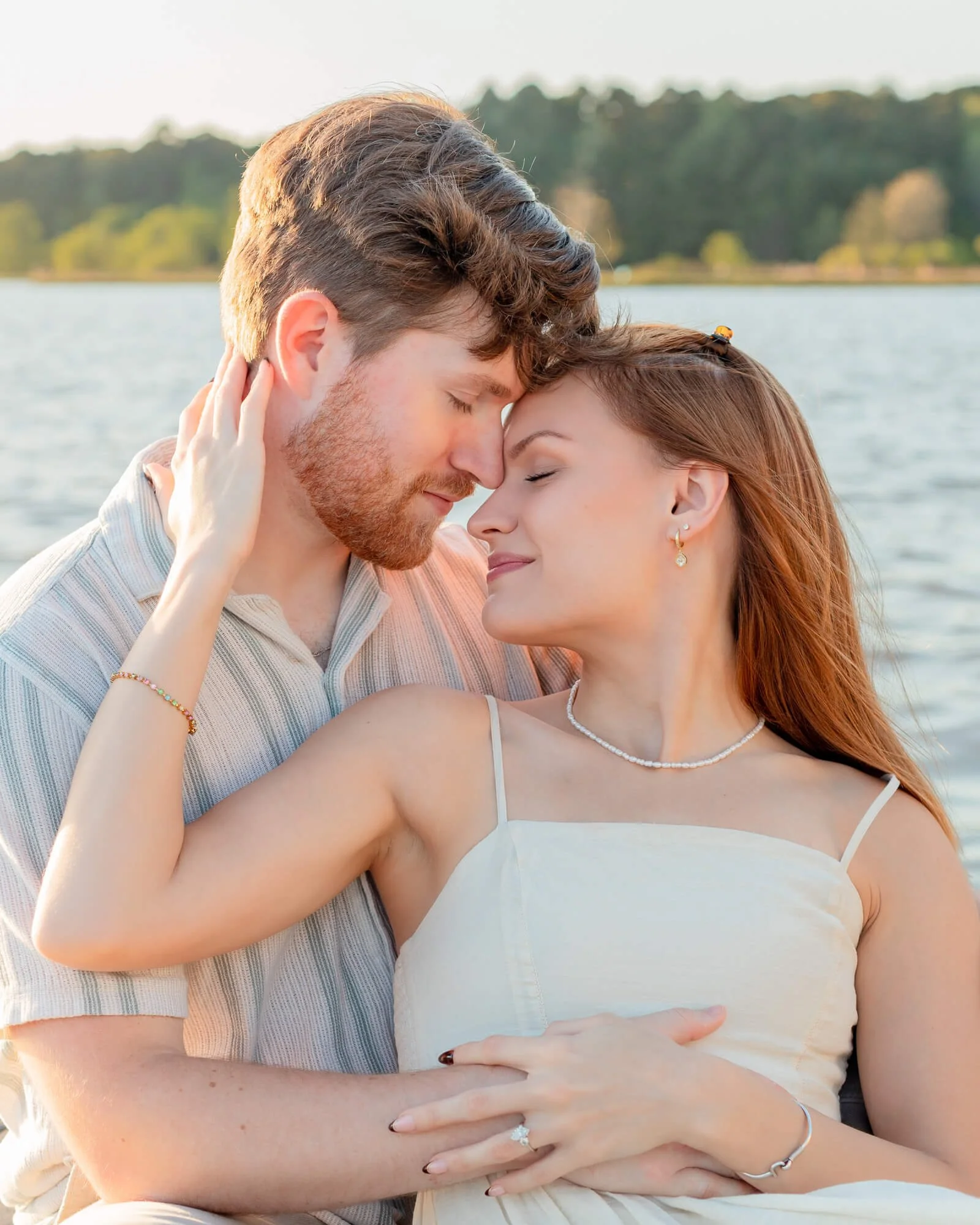 A man and woman embrace each other with their foreheads touching near a body of water with trees in the background, in a warm, outdoor setting.