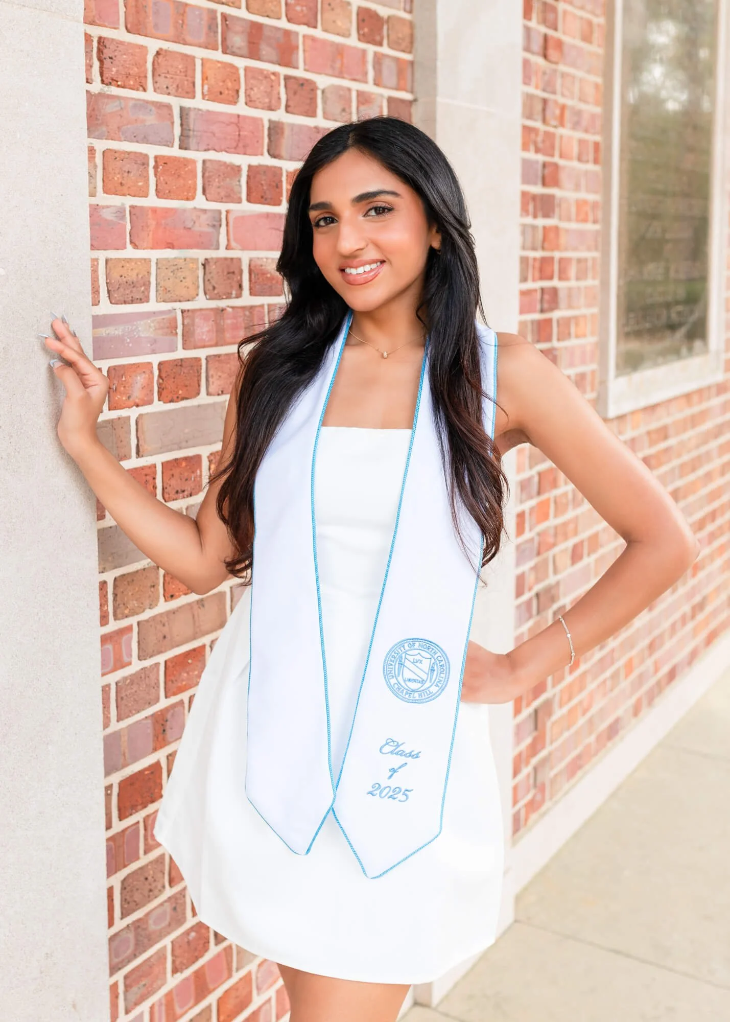 A young woman in a white dress and graduation stole, standing outside against a brick wall, smiling at the camera.