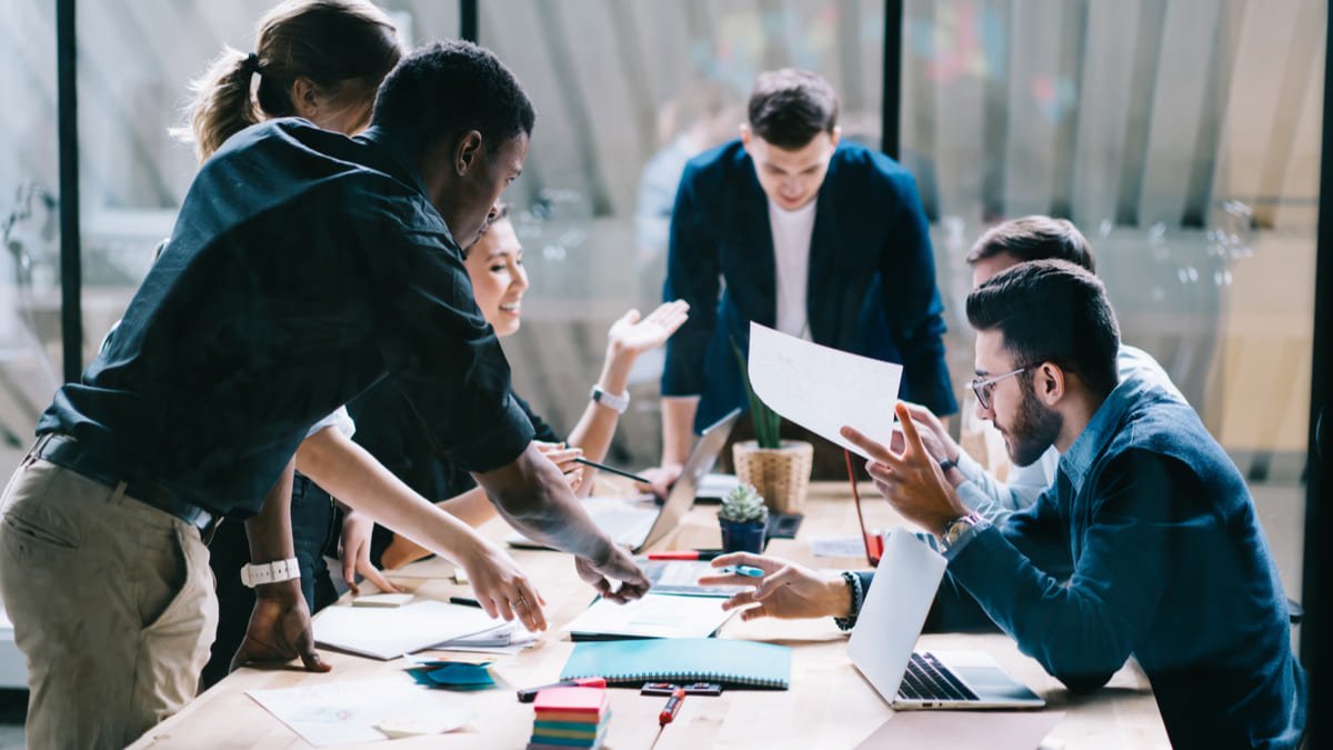 Group of diverse young professionals collaborating around a table in a modern office, discussing documents and working on laptops.