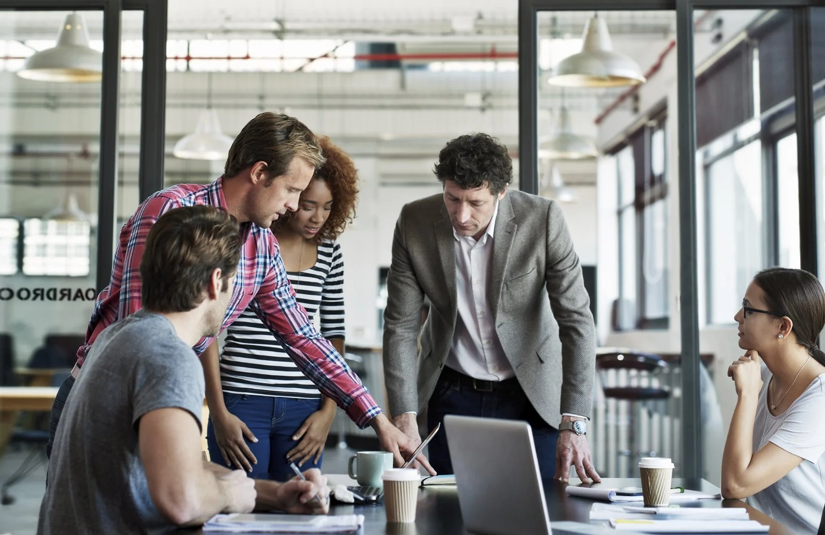 Group of five young professionals working together around a table in a modern office, looking at a tablet and documents.