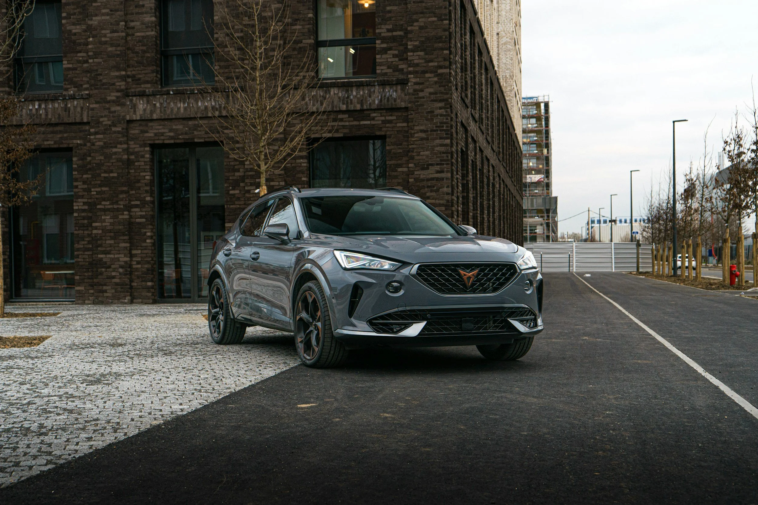 A gray modern compact SUV parked on a paved surface in front of a brick building with large windows, surrounded by trees and streetlights.