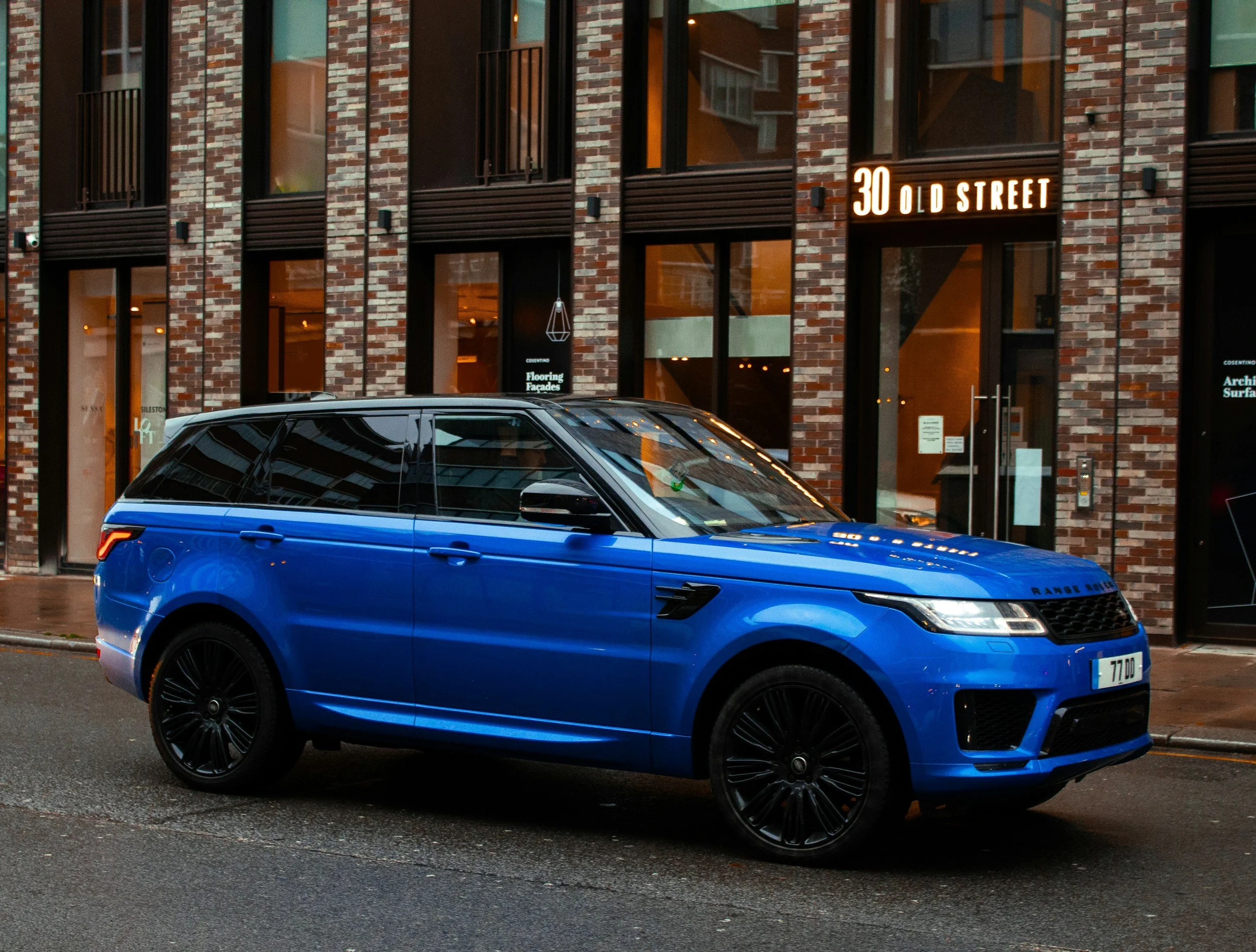 A blue Range Rover parked outside a brick building with large glass windows, on a city street at night.