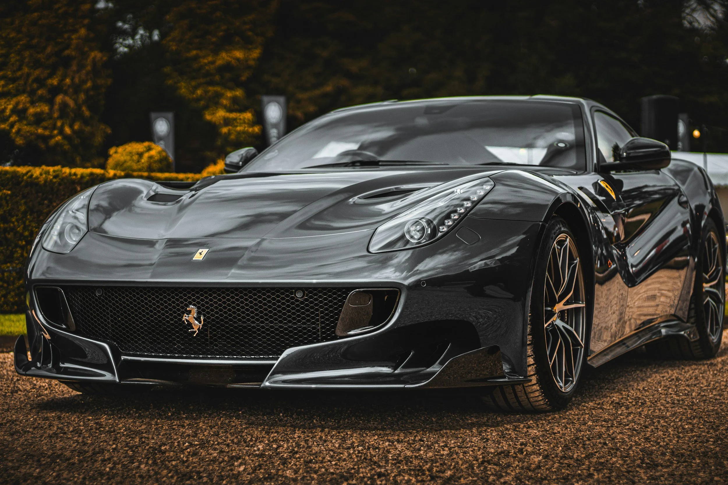 Black Ferrari sports car with sleek design and headlights, parked outdoors on a gravel surface with trees and banners in the background.