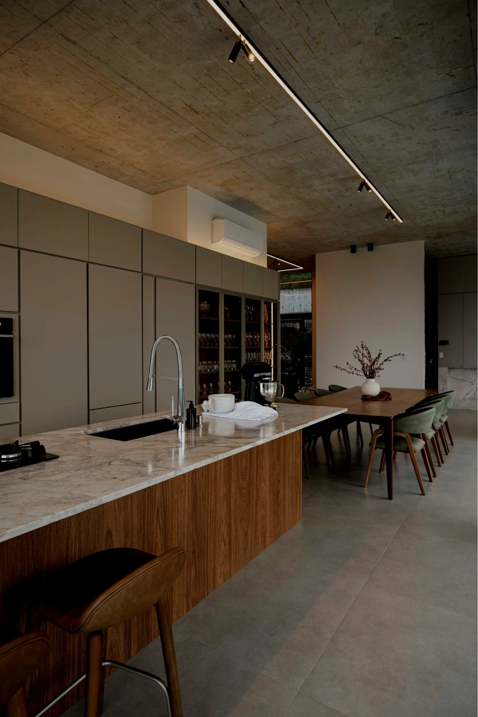 A neatly made dark blue upholstered bed with navy and white pillows, situated against a gray wall. Beside the bed is a modern white nightstand with two drawers, topped with a symmetric table lamp and a decorative black and white art piece. The room has a gray textured carpet.