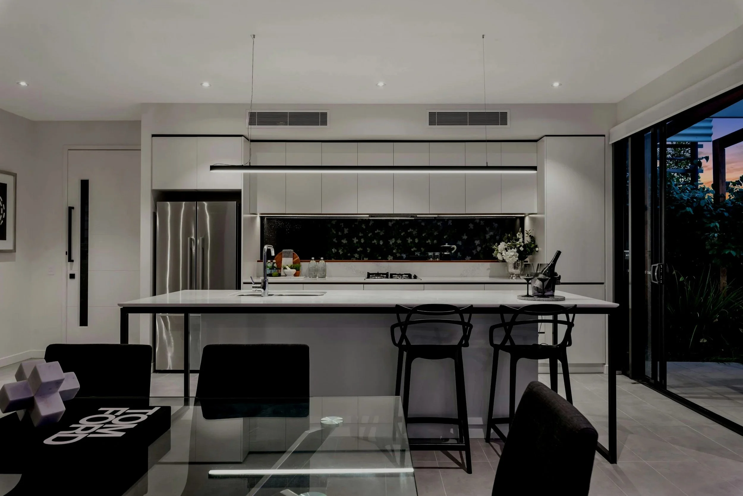 Living room with white walls, a large window, a white sofa with black and patterned pillows, a central coffee table with a large vase, two wooden armchairs with white cushions, a floor lamp, and a fireplace with a painting above it.