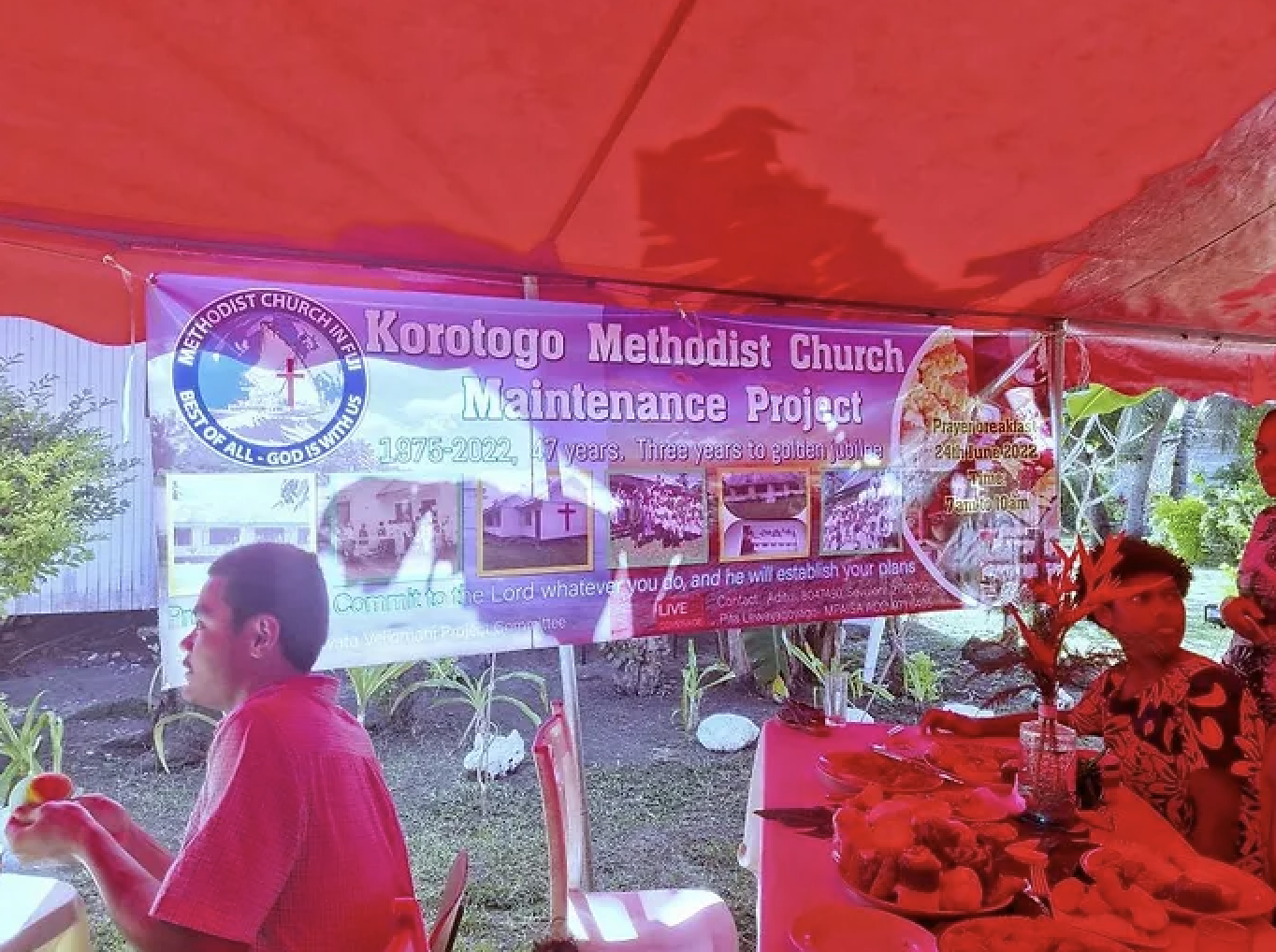 Banner for Korotogo Methodist Church Maintenance Project under a red tent with people seated at a table with food.