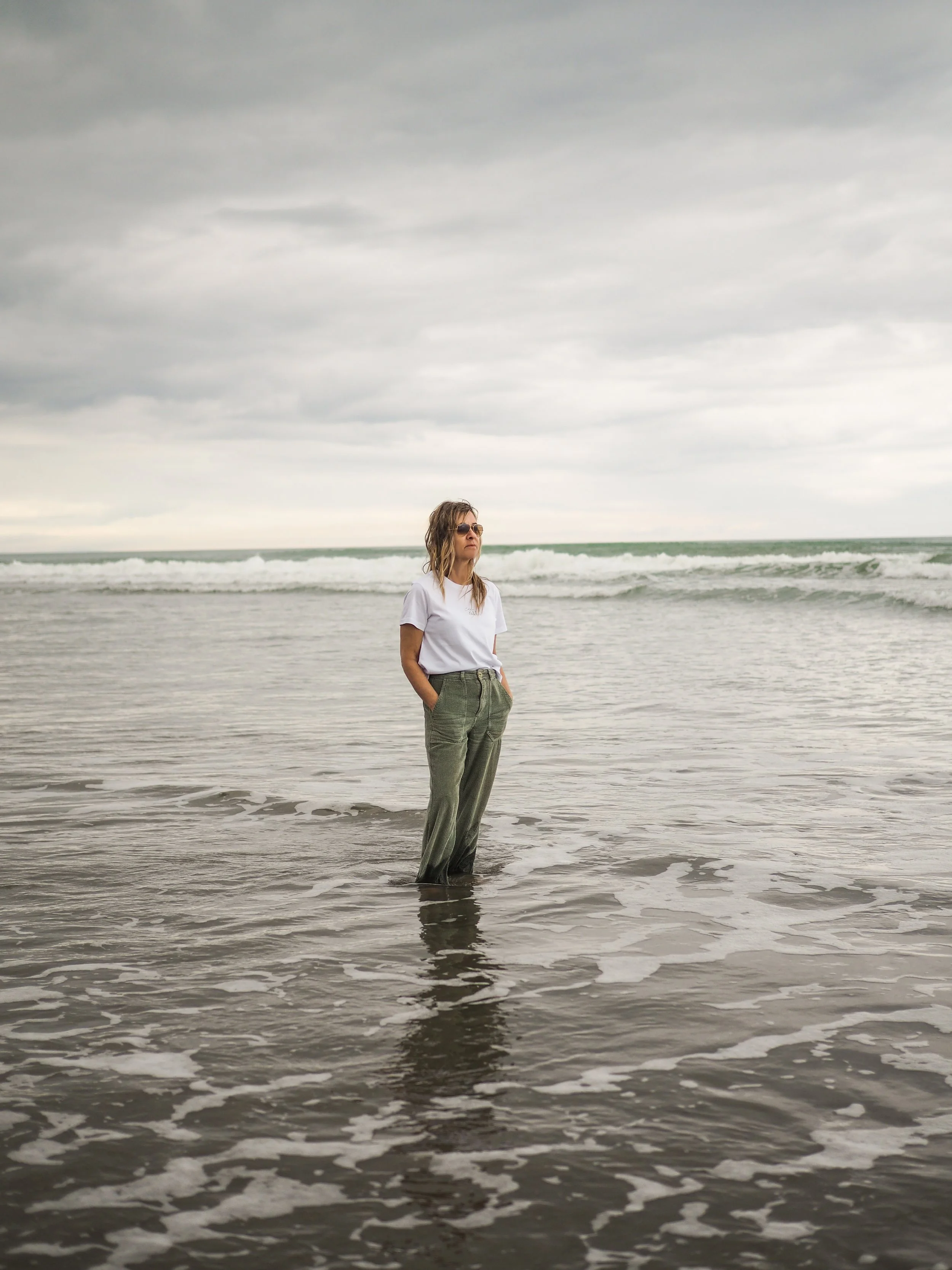 Person standing in shallow ocean water, wearing a white shirt and light green pants, overcast sky.