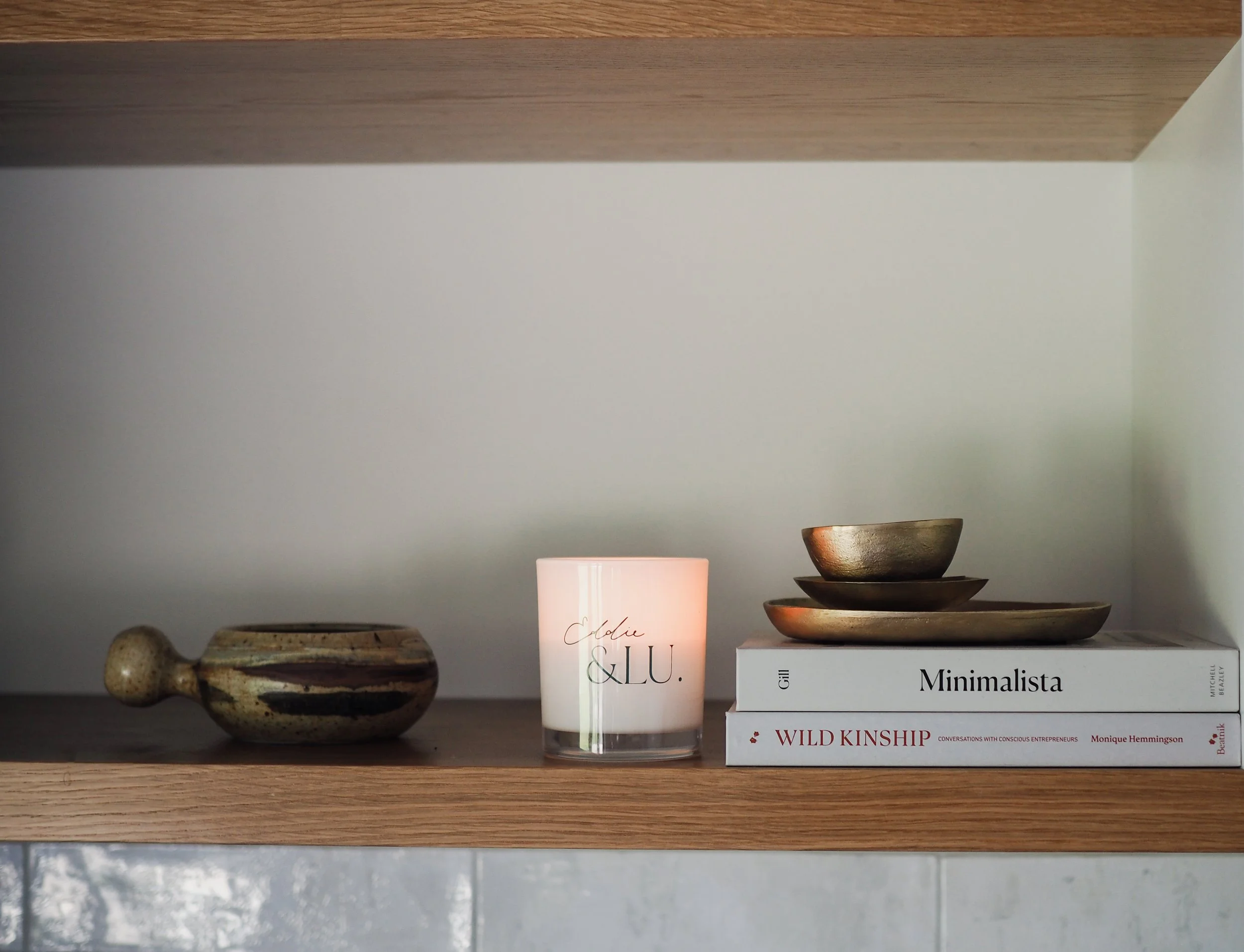 Wooden shelf displaying a ceramic bowl, a lit candle, two stacked books titled "Wild Kinship" and "Minimalista," and three nested bronze bowls.