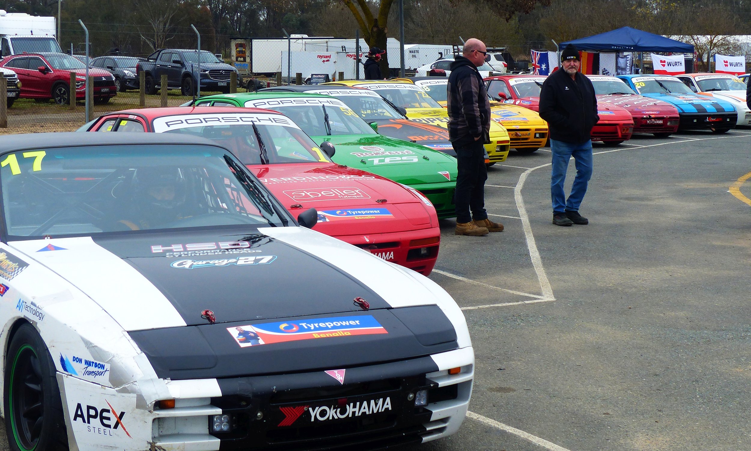 Race cars parked in a line at a racing event, with two men standing nearby, one wearing sunglasses and a black jacket, the other in a hoodie. The cars are colorful, with branding and sponsorship logos, and a fenced area with other vehicles and flags can be seen in the background.