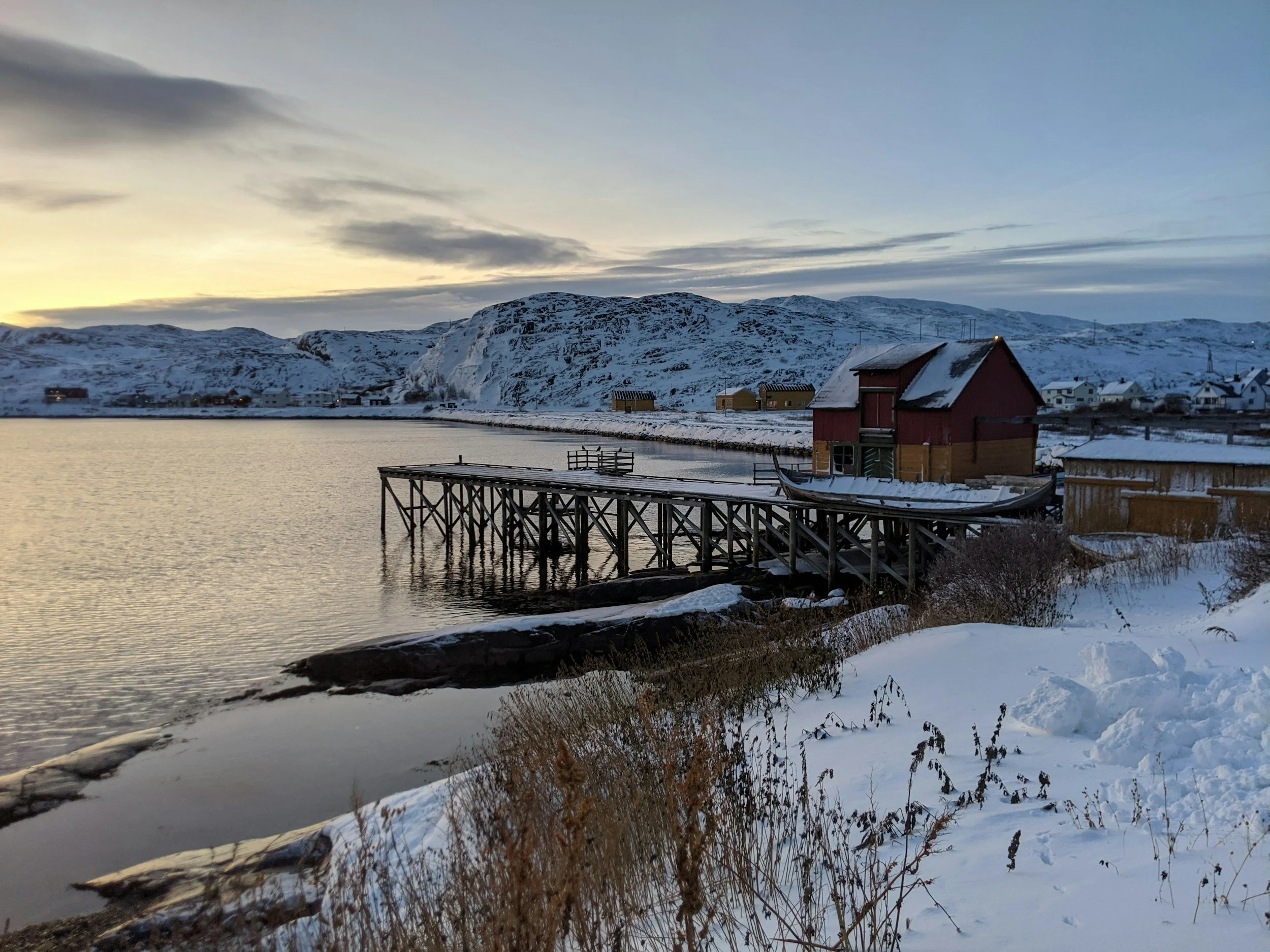 Snowy landscape with wooden pier and red building next to calm water, surrounded by snow-covered mountains during sunset.