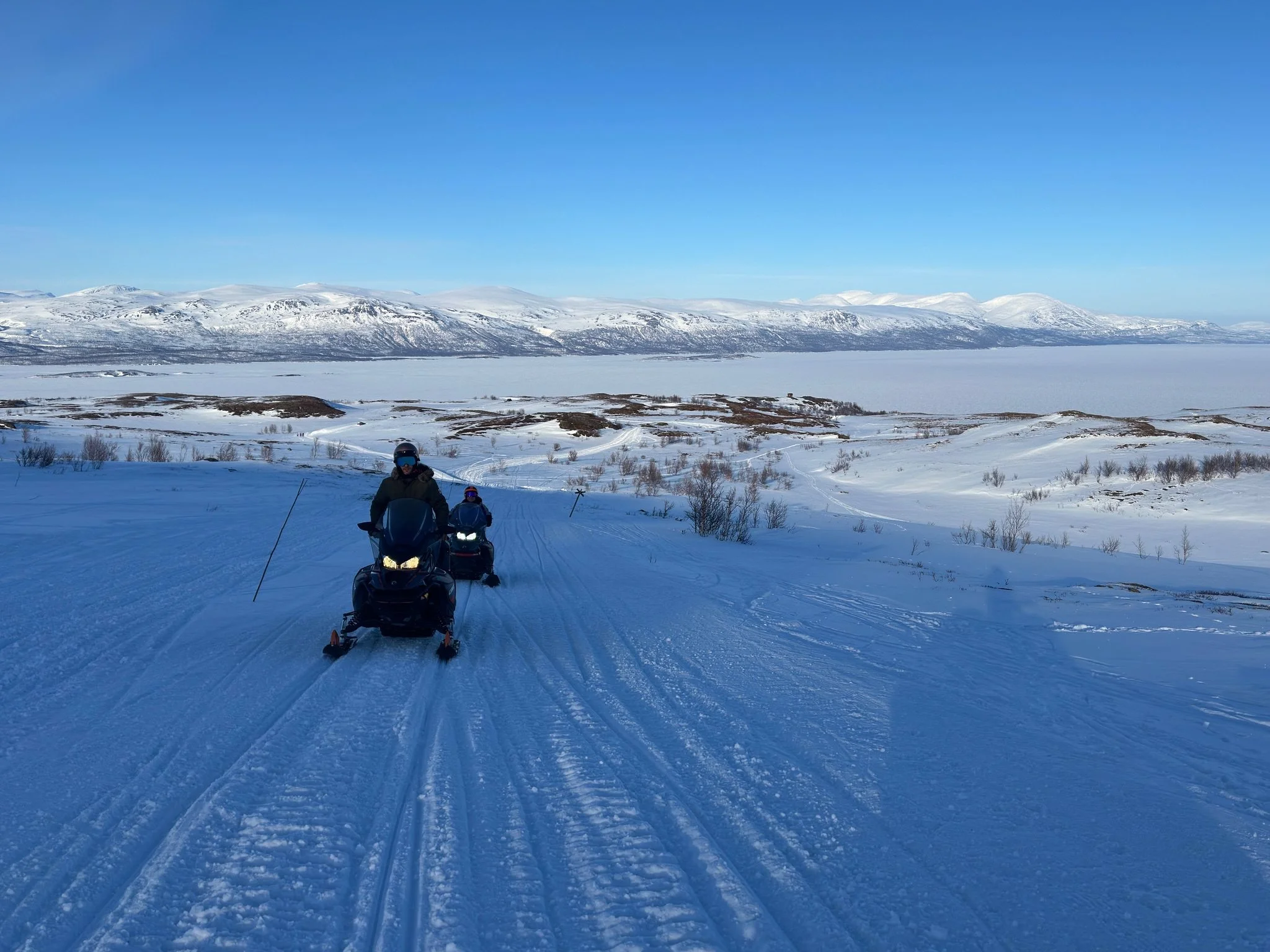 Two people riding snowmobiles on a snowy trail with a backdrop of snow-covered mountains and a clear blue sky.