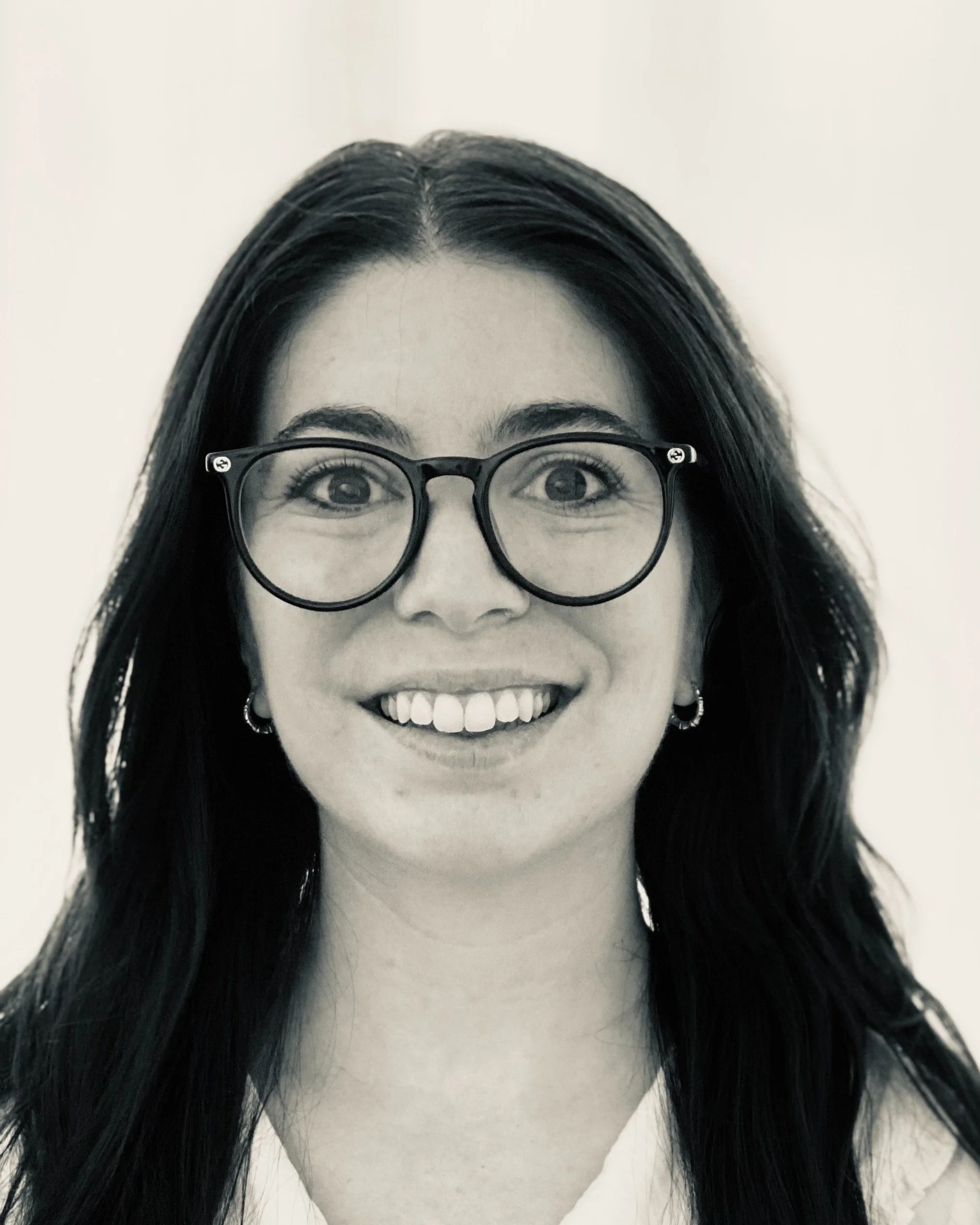 Black-and-white photo of a woman with long dark hair, glasses, earrings, and a big smile.