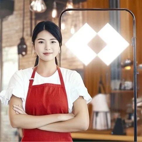 Young woman in a white shirt and red apron standing with arms crossed inside a restaurant, with warm lighting and wooden decor, and a large illuminated Techcombank logo light in the background.