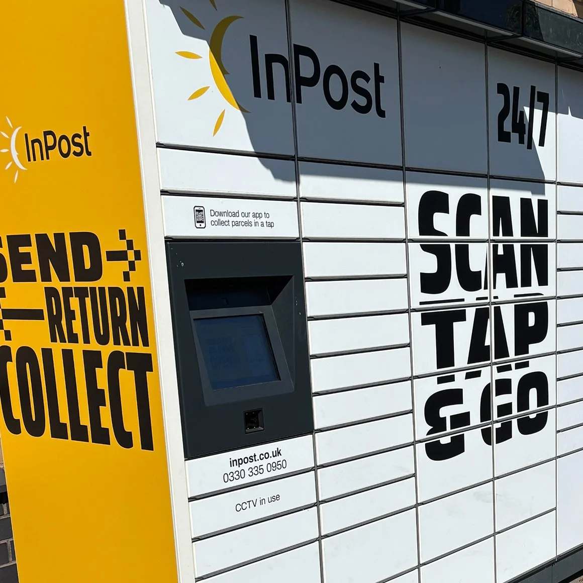 A parcel locker with a yellow sign on the side that reads 'Send', 'Return', 'Collect', and instructions to download an app. The lockers are labeled with the words 'InPost' and 'Scan Tap' along with the hours 24/7. A screen and barcode scanner are on the locker.