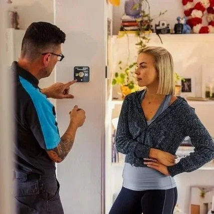 An engineer fits a thermostat takes with a woman indoors, with Christmas decorations in the background.