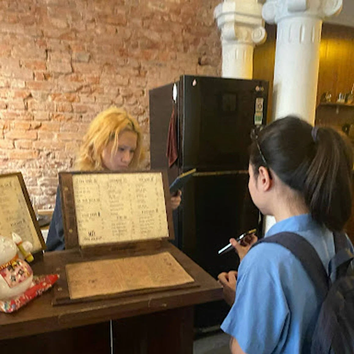 A woman and a girl look at a restaurant menu in a brick-walled restaurant with columns and a refrigerator.