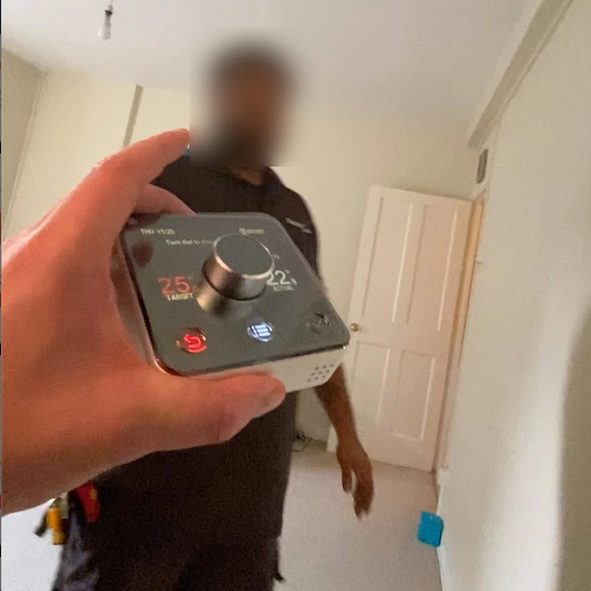 Person holding a thermostat in front of a mirror, reflecting a blurred man standing in a room with a white door.