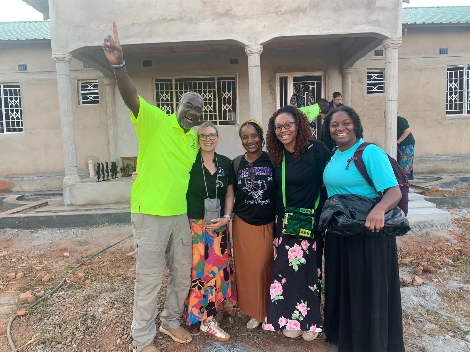 Group of five smiling people standing in front of a partially built house, posing for a photo, with construction materials on the ground.