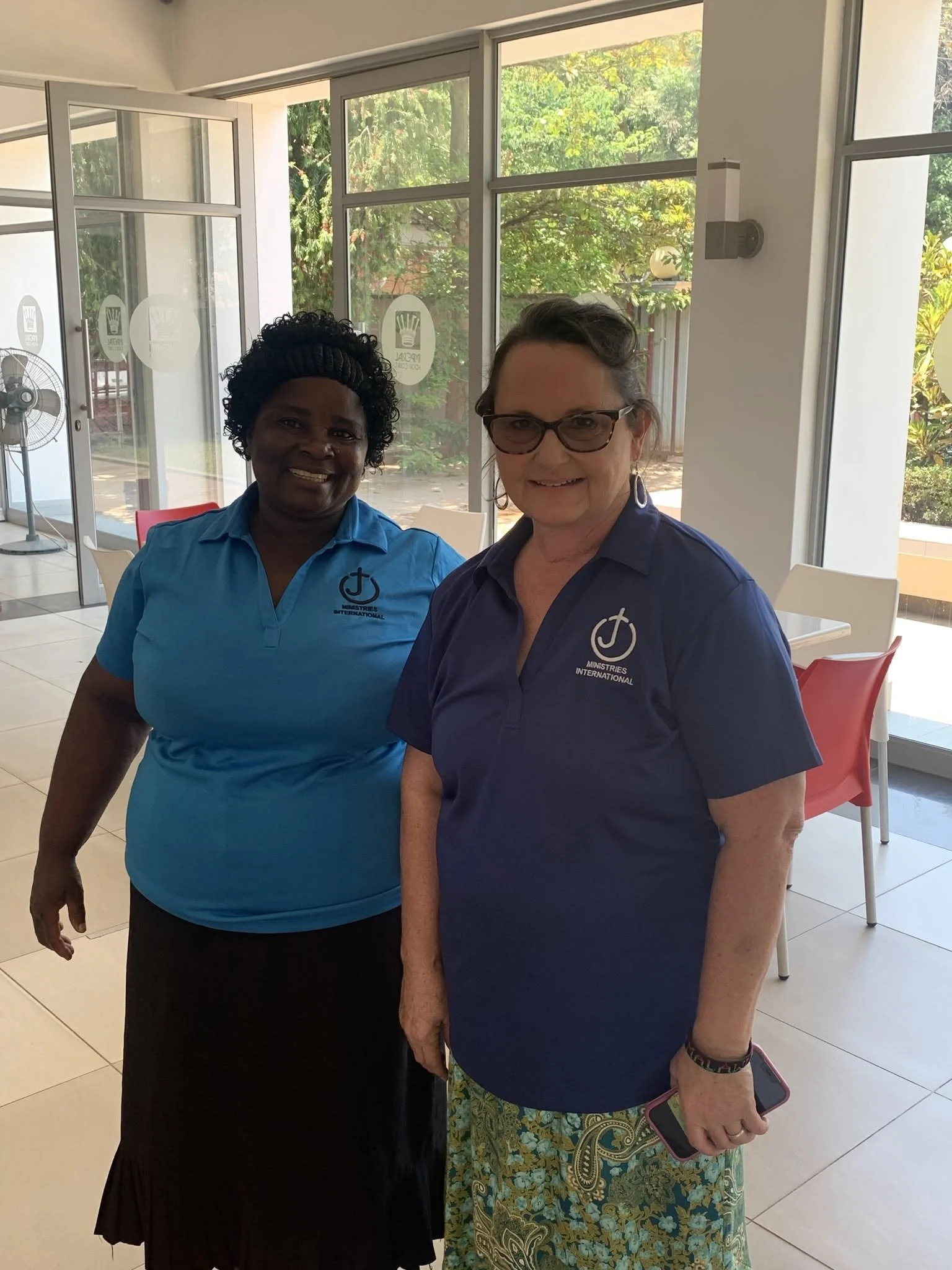 Two women standing indoors near large windows with greenery outside, smiling at the camera. One is wearing a blue shirt with a Ministries International logo, and the other is wearing a dark blue shirt with a similar logo, holding a phone and wearing glasses.