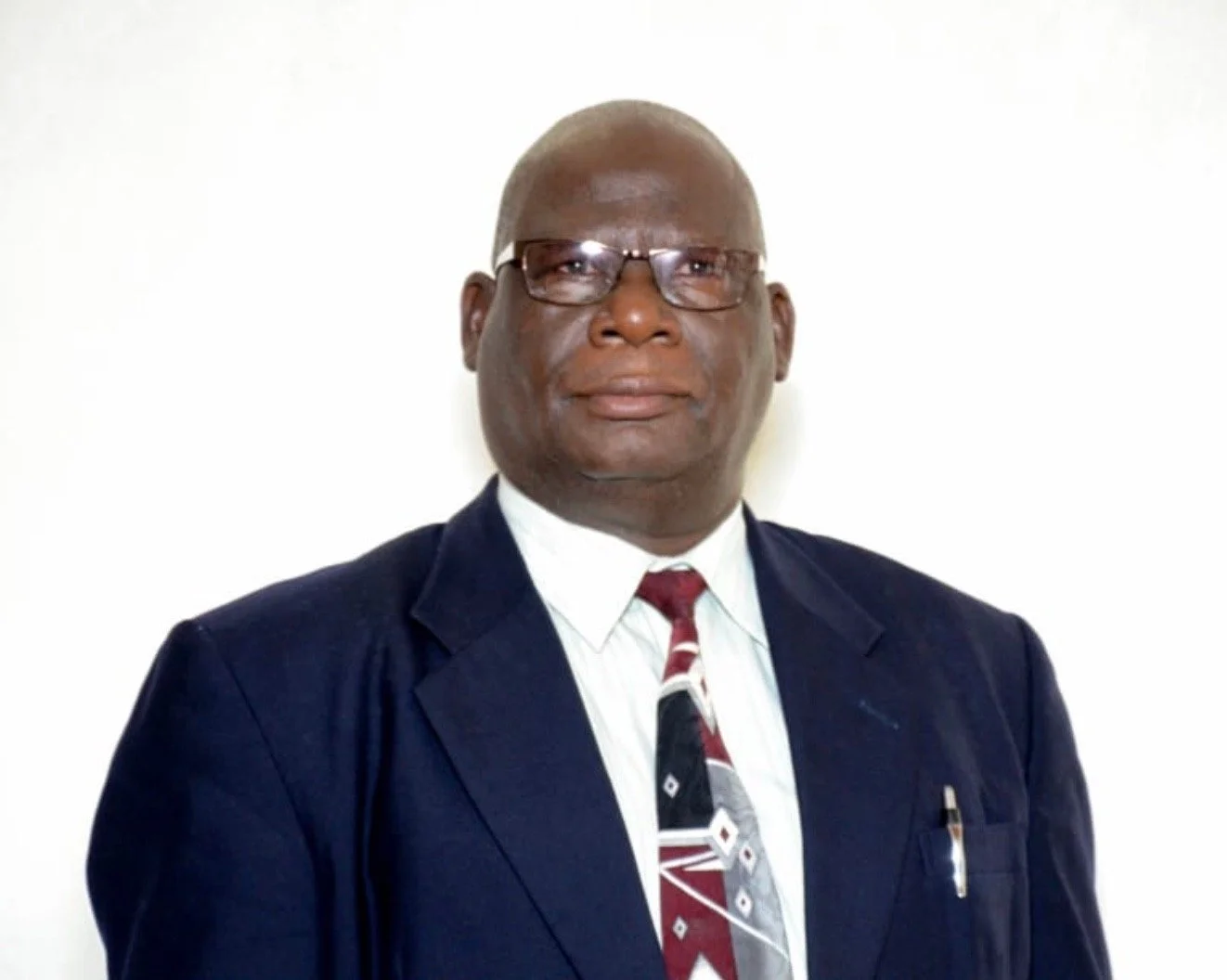 A man wearing glasses, dressed in a navy suit, white shirt, and patterned tie, stands against a white background.