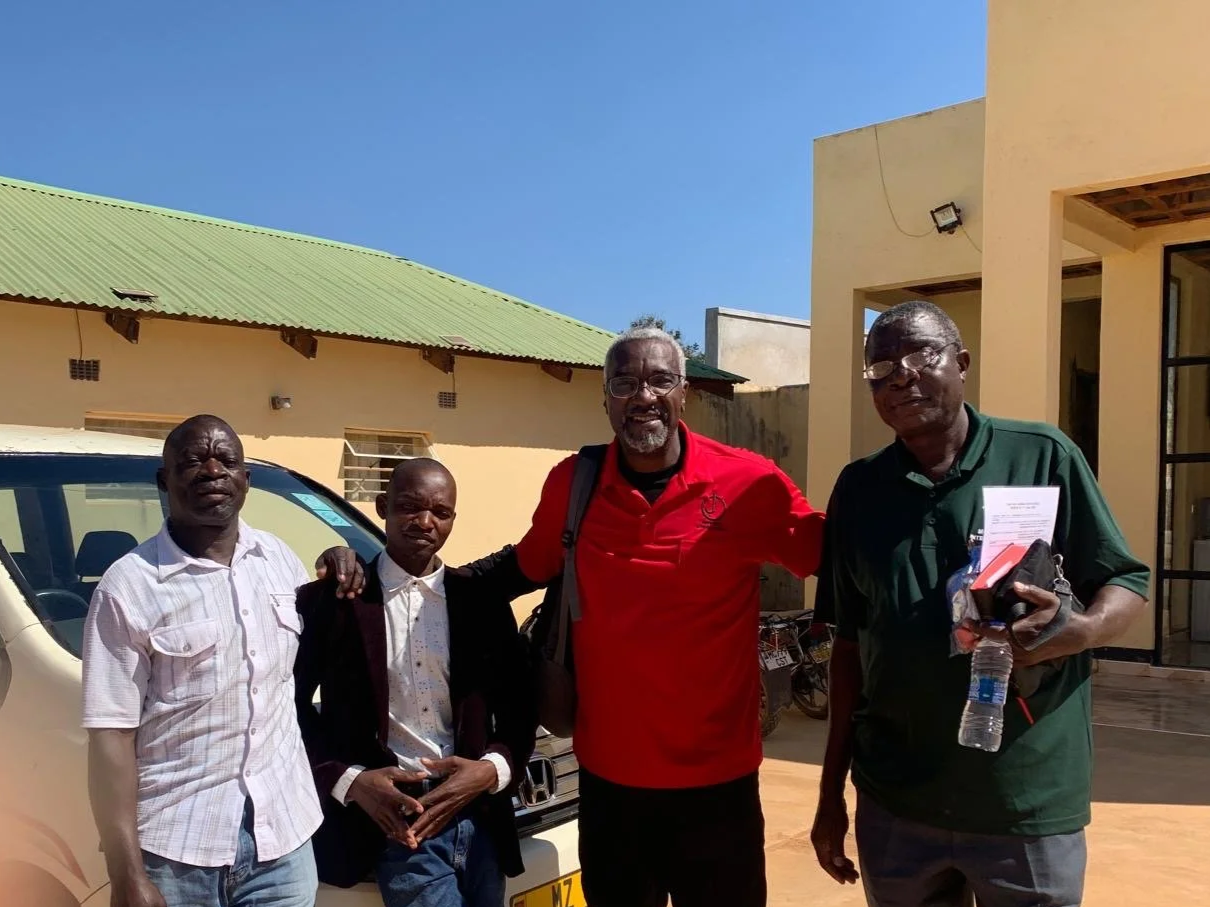 Four men standing outside in front of a yellow building and a vehicle under a blue sky.