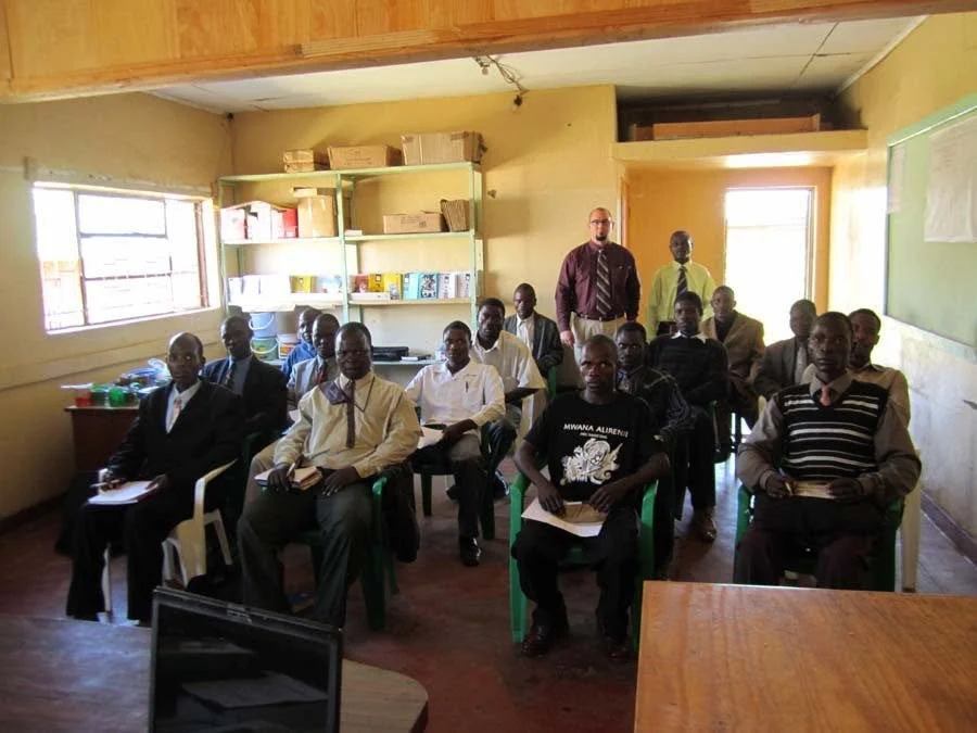A classroom with students and two teachers, some students are holding notebooks, shelves with books and supplies, windows providing natural light, and a chalkboard on the wall.