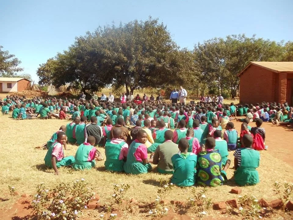 Large group of school children seated outdoors on grass, wearing school uniforms, listening to presenters near trees and school buildings.