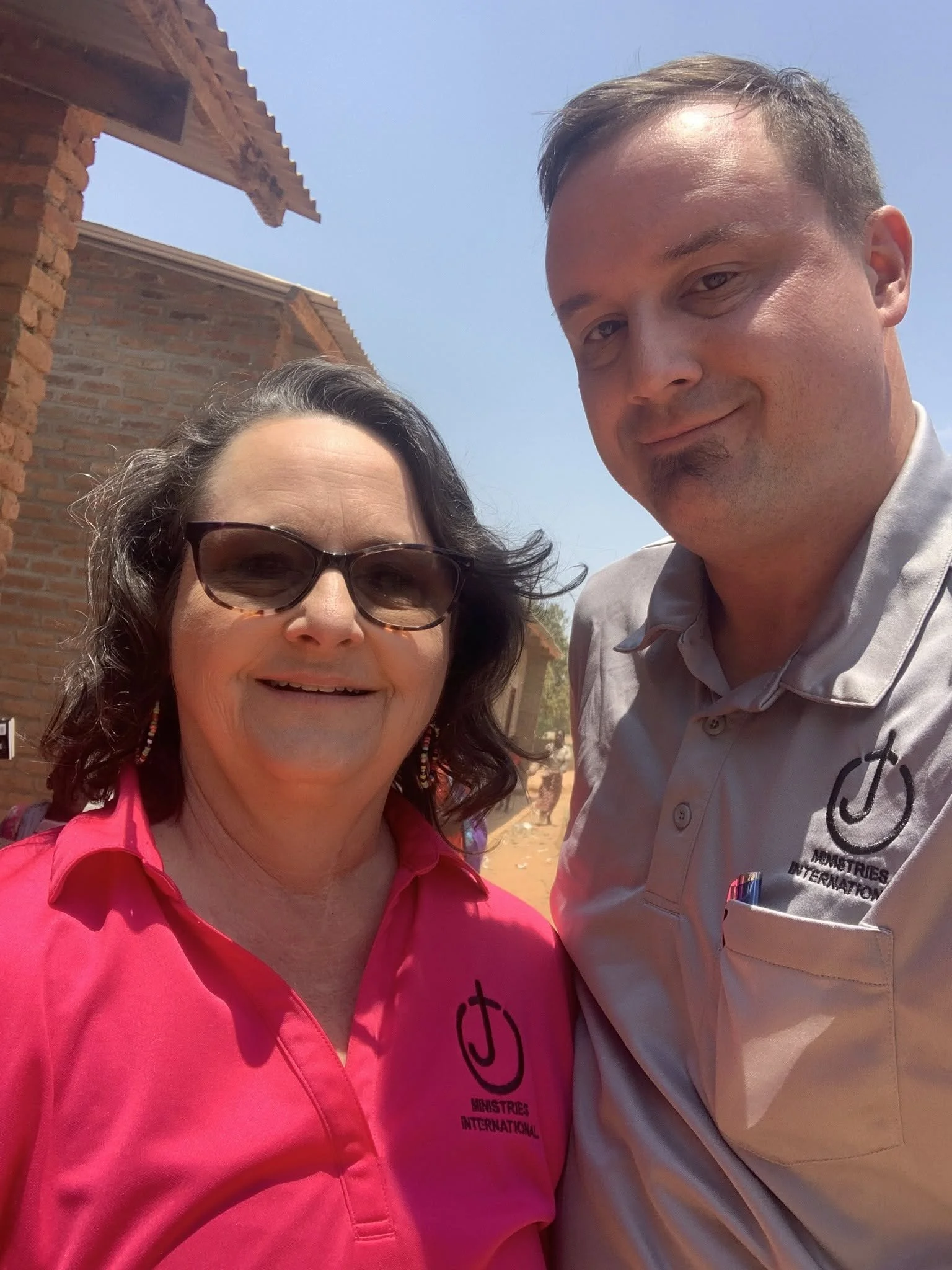 A woman wearing sunglasses and a pink shirt, and a man wearing a beige shirt with a logo, taking a selfie outdoors with a clear blue sky and some brick structures in the background.